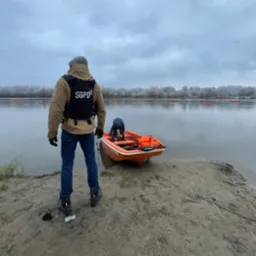 Funkcjonariusz SGPD w kamizelce taktycznej stoi tyłem, obserwując pracę kolegi przy pomarańczowej łodzi na brzegu rzeki, na tle zalesionego brzegu i pochmurnego nieba.