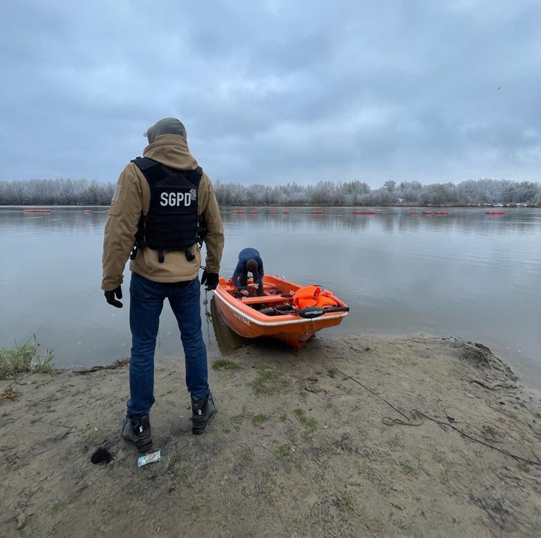 Funkcjonariusz SGPD w kamizelce taktycznej stoi tyłem, obserwując pracę kolegi przy pomarańczowej łodzi na brzegu rzeki, na tle zalesionego brzegu i pochmurnego nieba.