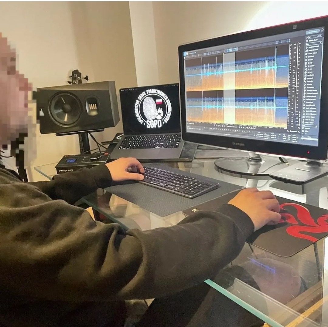 Osoba pracująca przy biurku z dwoma monitorami, laptopem z logo SGPD, klawiaturą i myszką, analizująca dane graficzne na ekranie, z widocznym głośnikiem studyjnym.