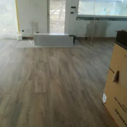 Wnętrze w trakcie remontu: ułożone panele podłogowe o wzorze drewna, przygotowane płyty do montażu, zakryte okno folią ochronną, widoczne przewody elektryczne i elementy instalacji.