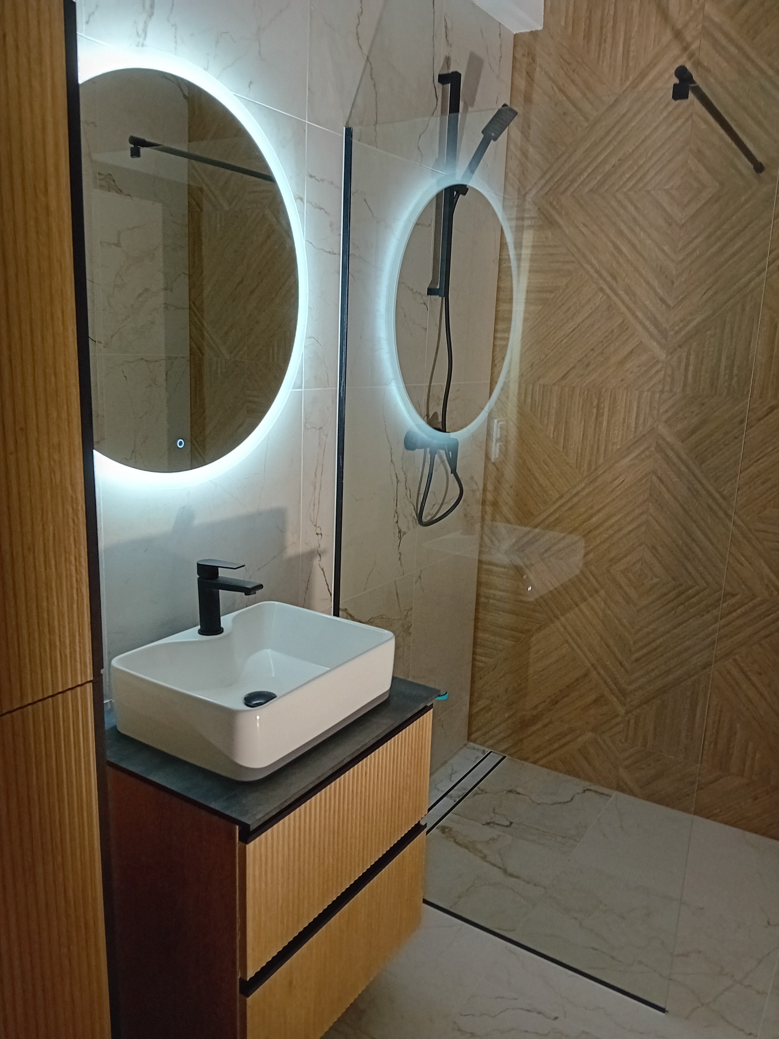 Nowoczesna łazienka z umywalką nablatową, podświetlanym lustrem i kabiną prysznicową ze szklaną ścianką oraz geometrycznymi płytkami w odcieniach beżu.