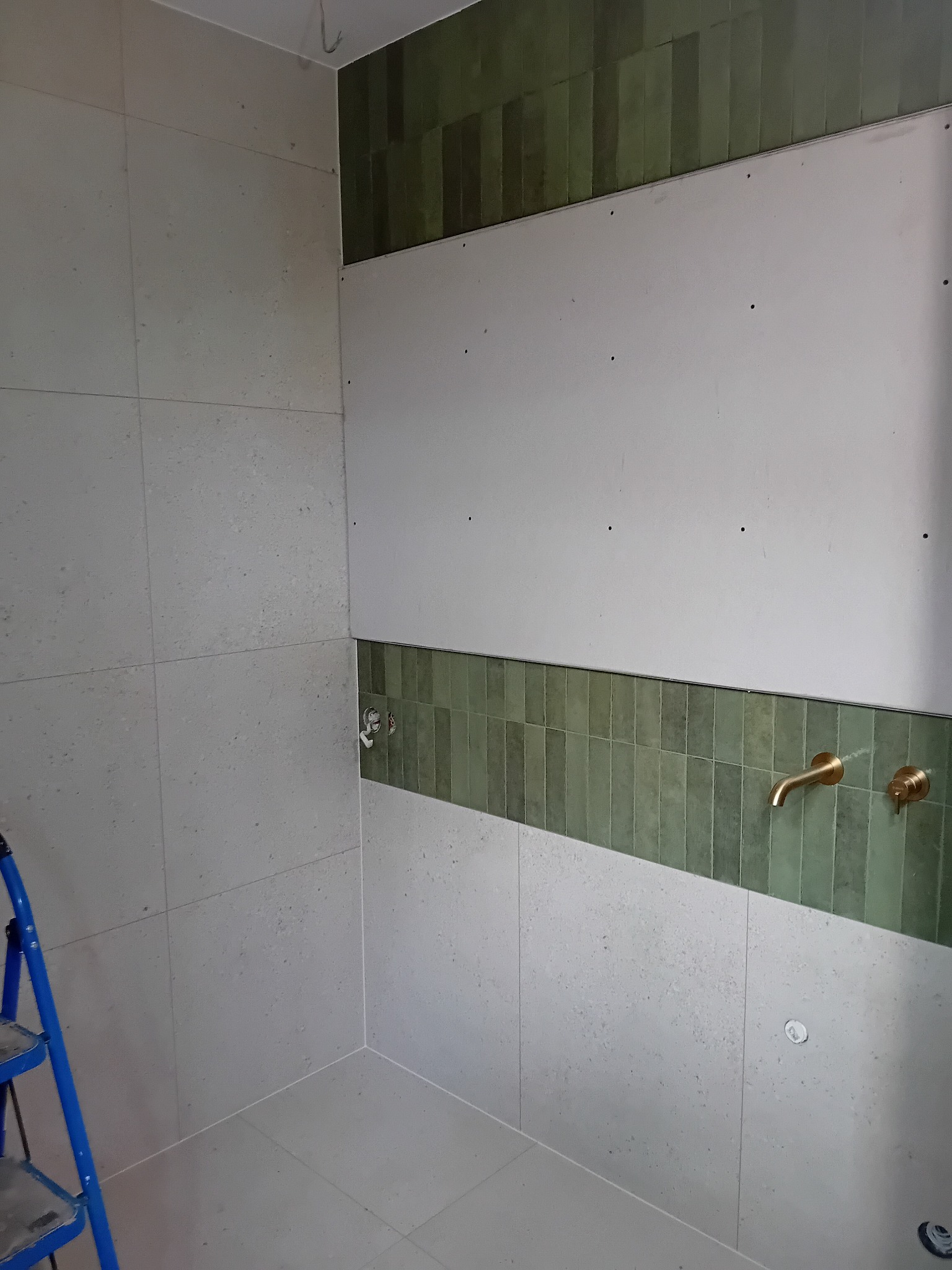 Fragment łazienki w trakcie remontu: ułożone szare płytki na ścianach, pas zielonych, pionowych płytek dekoracyjnych, surowa ściana z płyt gipsowo-kartonowych, zamontowane złote baterie umywalkowe,...