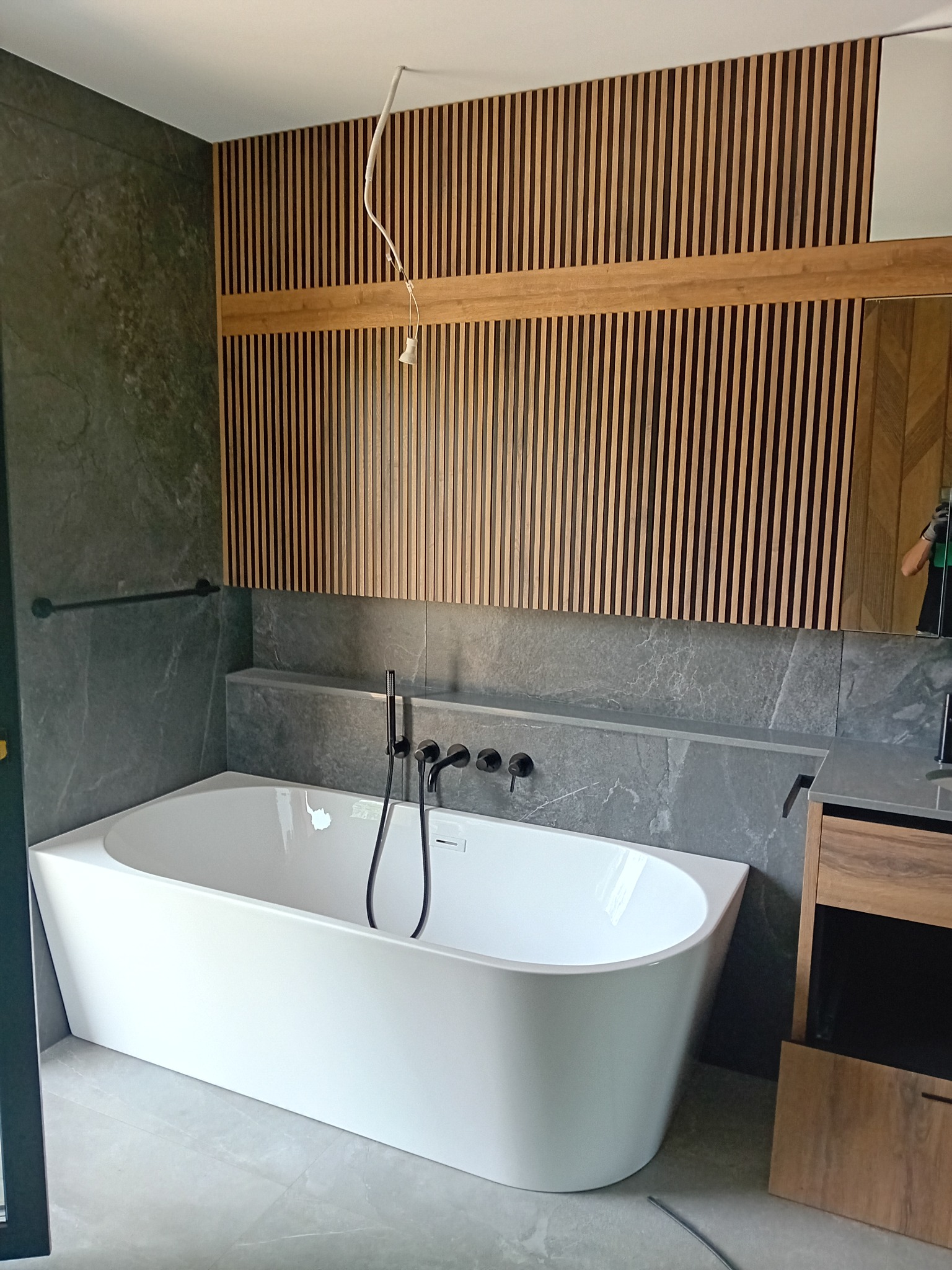 Nowoczesna łazienka z białą wanną wolnostojącą, szarą płytką imitującą kamień i drewnianymi lamelami na ścianie, czarna armatura, niedokończone podłączenie oświetlenia.