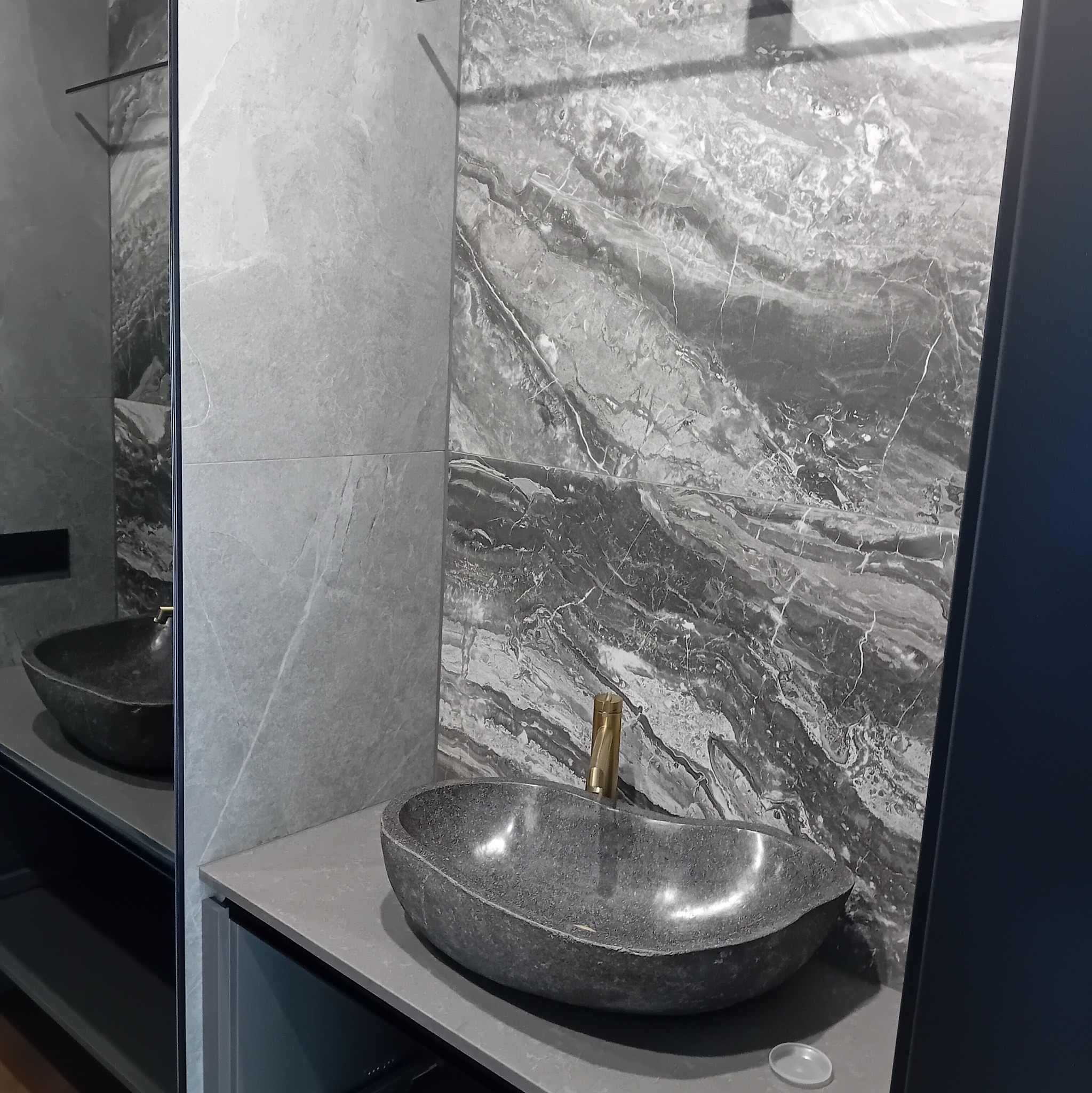 Nowoczesna łazienka z umywalką z szarego kamienia i złotą baterią na tle płytek imitujących marmur.