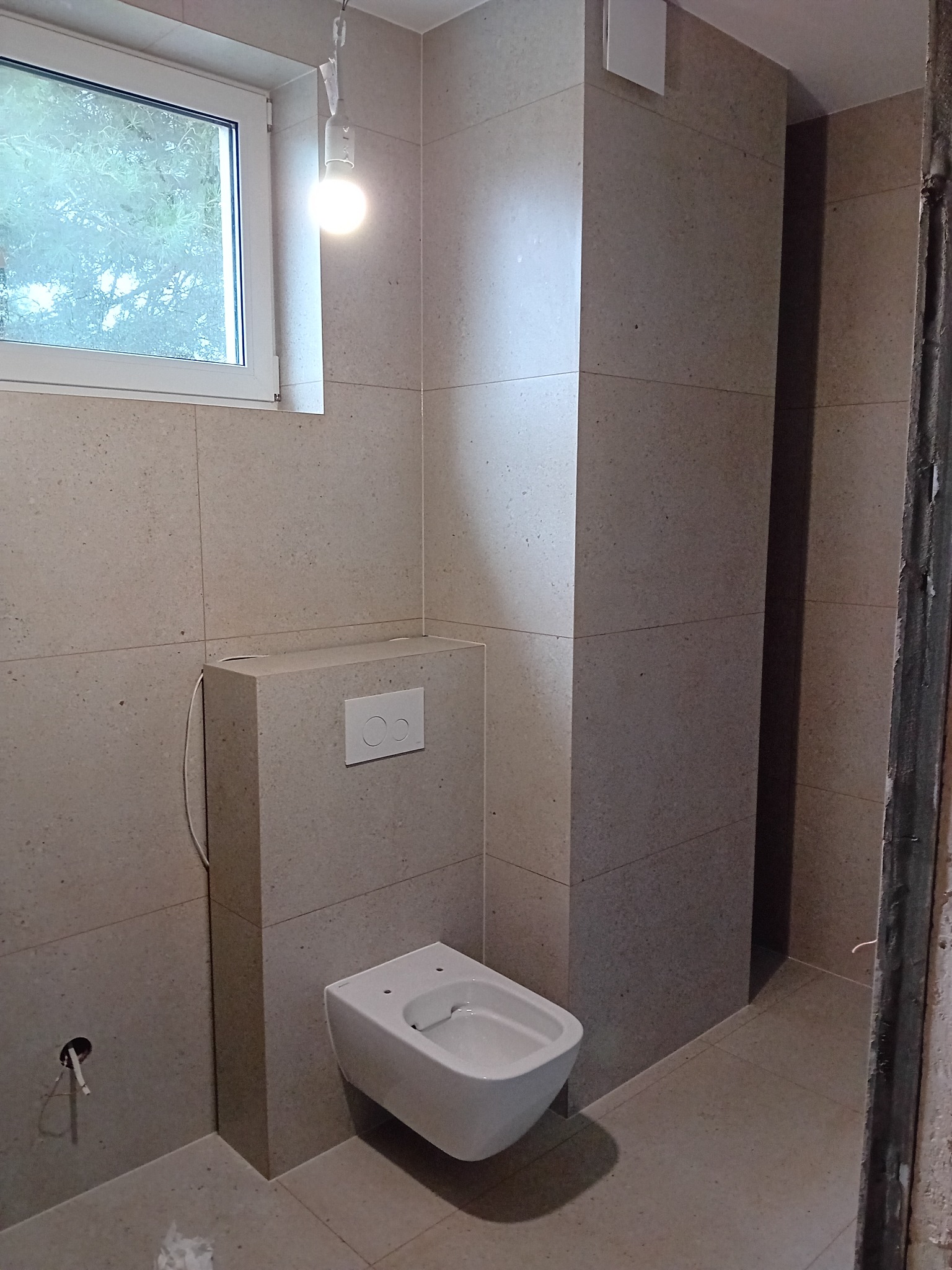 Ściana i podłoga w łazience wyłożone jasnymi płytkami imitującymi kamień, wisząca toaleta z podtynkową spłuczką, widoczny otwór na rurę, surowa ościeżnica i tymczasowa żarówka.