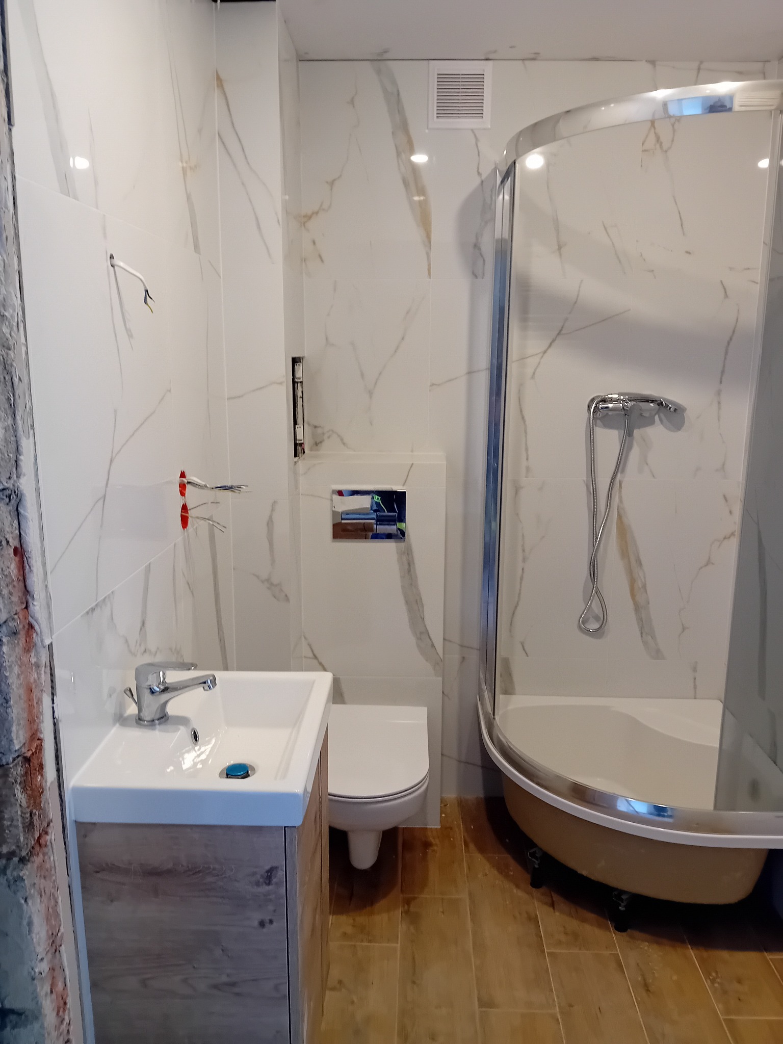 Wykończona łazienka z kabiną prysznicową, umywalką z szafką imitującą drewno oraz toaletą, białe płytki imitujące marmur na ścianach, brązowe płytki na podłodze, widoczne wystające kable elektryczne.