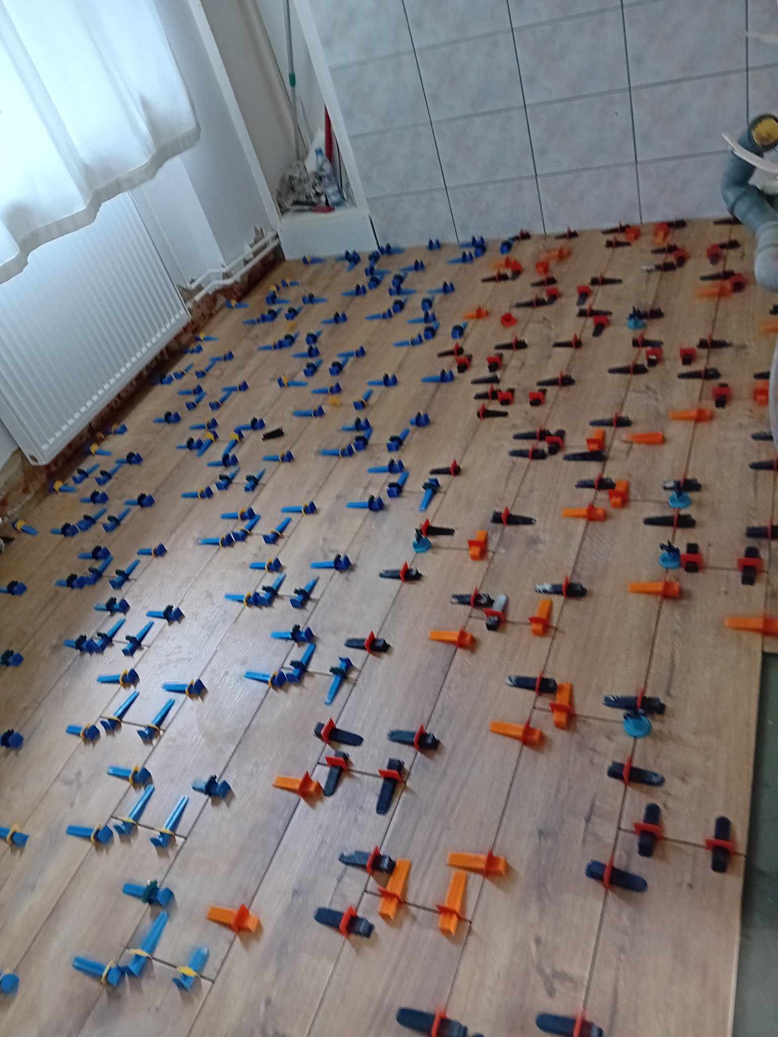 Proces układania płytek imitujących drewno z użyciem poziomujących klipsów i klinów w kolorach niebieskim, pomarańczowym i czarnym; widoczna część łazienki z grzejnikiem i fragmentem ściany...