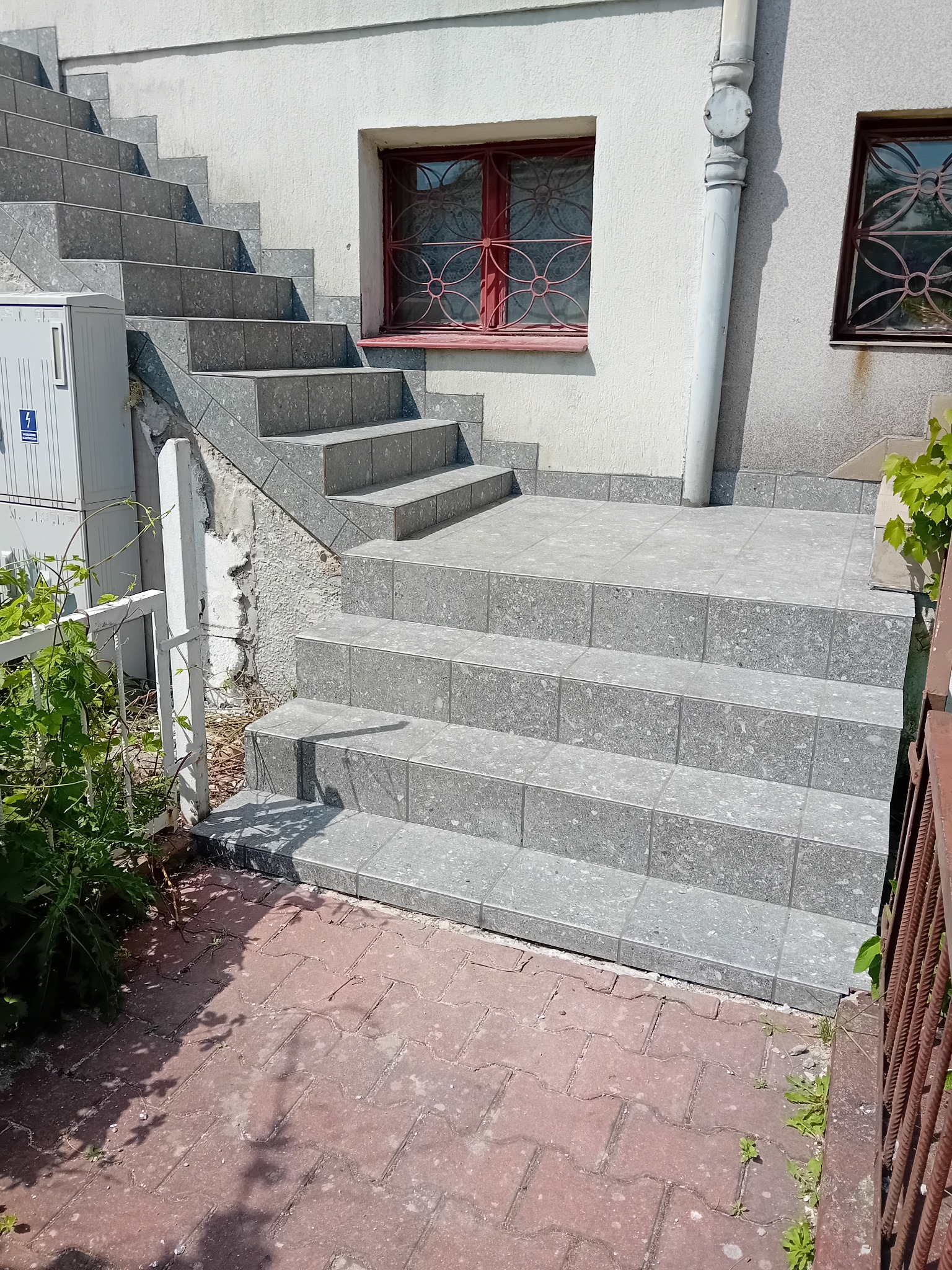Zewnętrzne schody wejściowe wyłożone szarymi płytkami z ciemniejszymi plamkami, prowadzące do drzwi z ozdobną kratą, na tle białej ściany budynku.