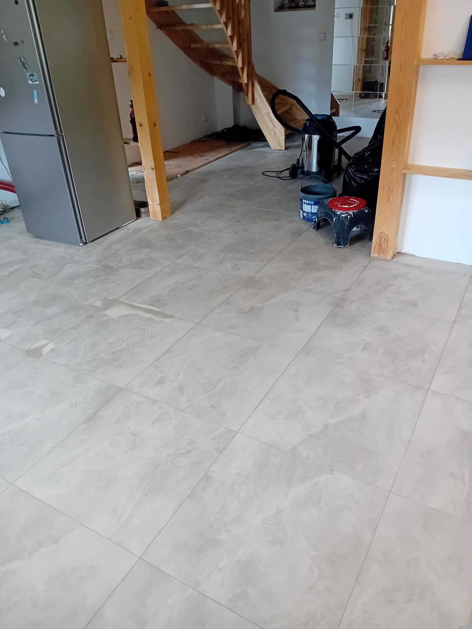 Jasne, marmurkowe płytki podłogowe ułożone w nowym domu, widoczne ślady po budowie, schody drewniane w tle, lodówka i narzędzia glazurnicze.