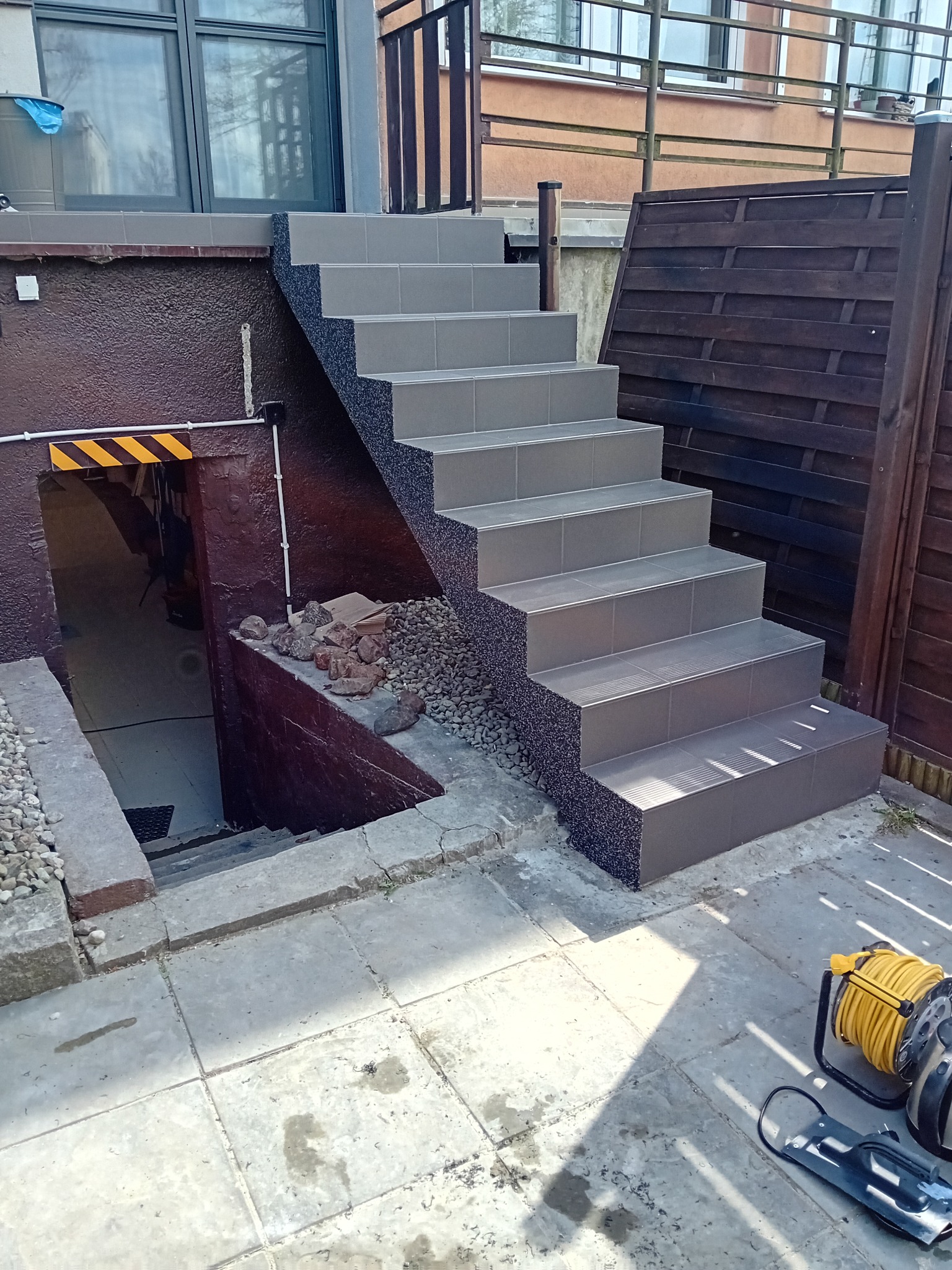 Zewnętrzne schody wyłożone szarymi płytkami prowadzące do piwnicy, z widocznym kablem elektrycznym i fragmentem drewnianego ogrodzenia.