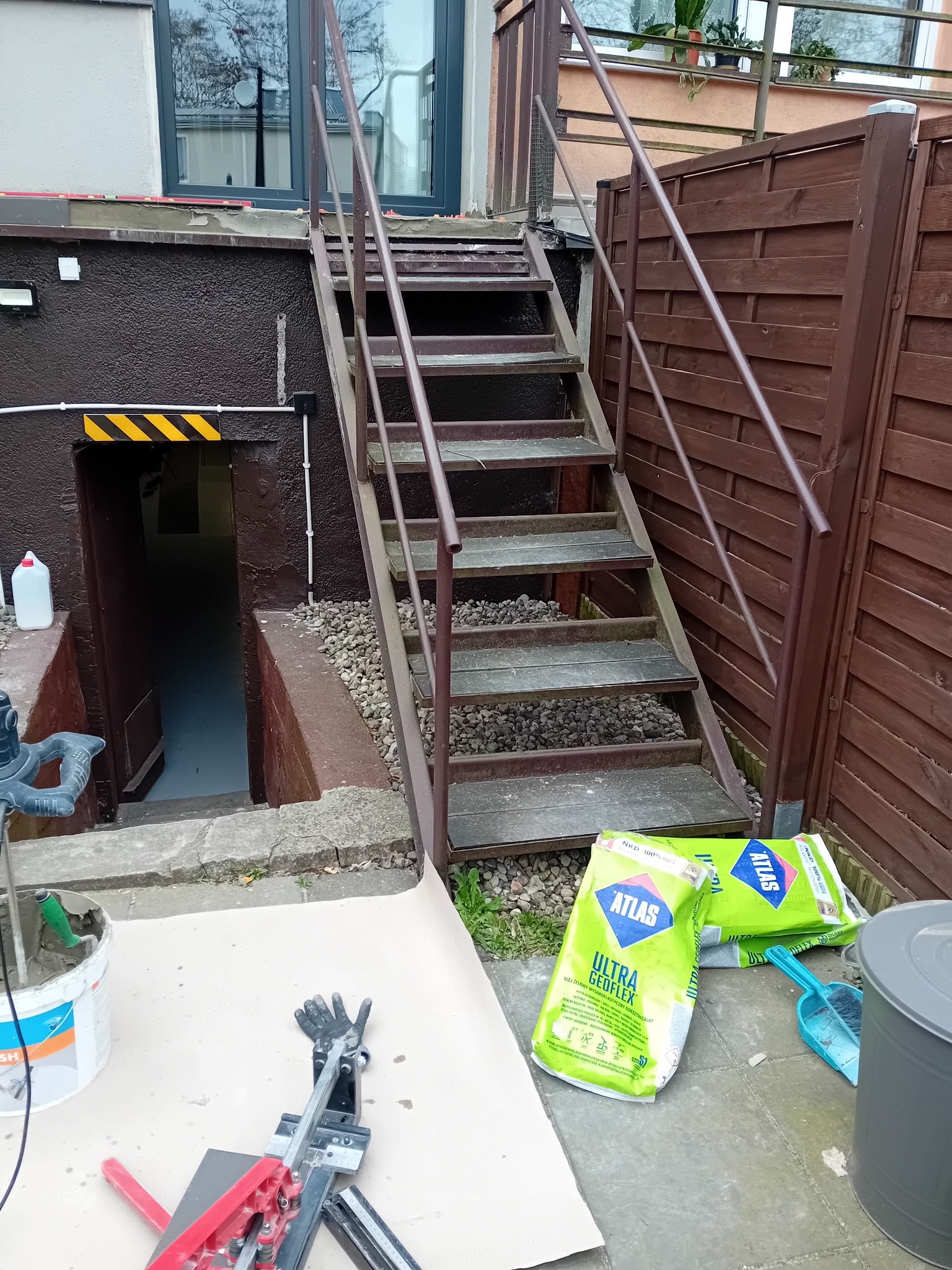 Przygotowane narzędzia i materiały budowlane, w tym wiadro z zaprawą, przecinarka do płytek i worki z klejem Atlas Ultra Geoflex, ułożone na zewnątrz budynku, obok metalowych schodów prowadzących...