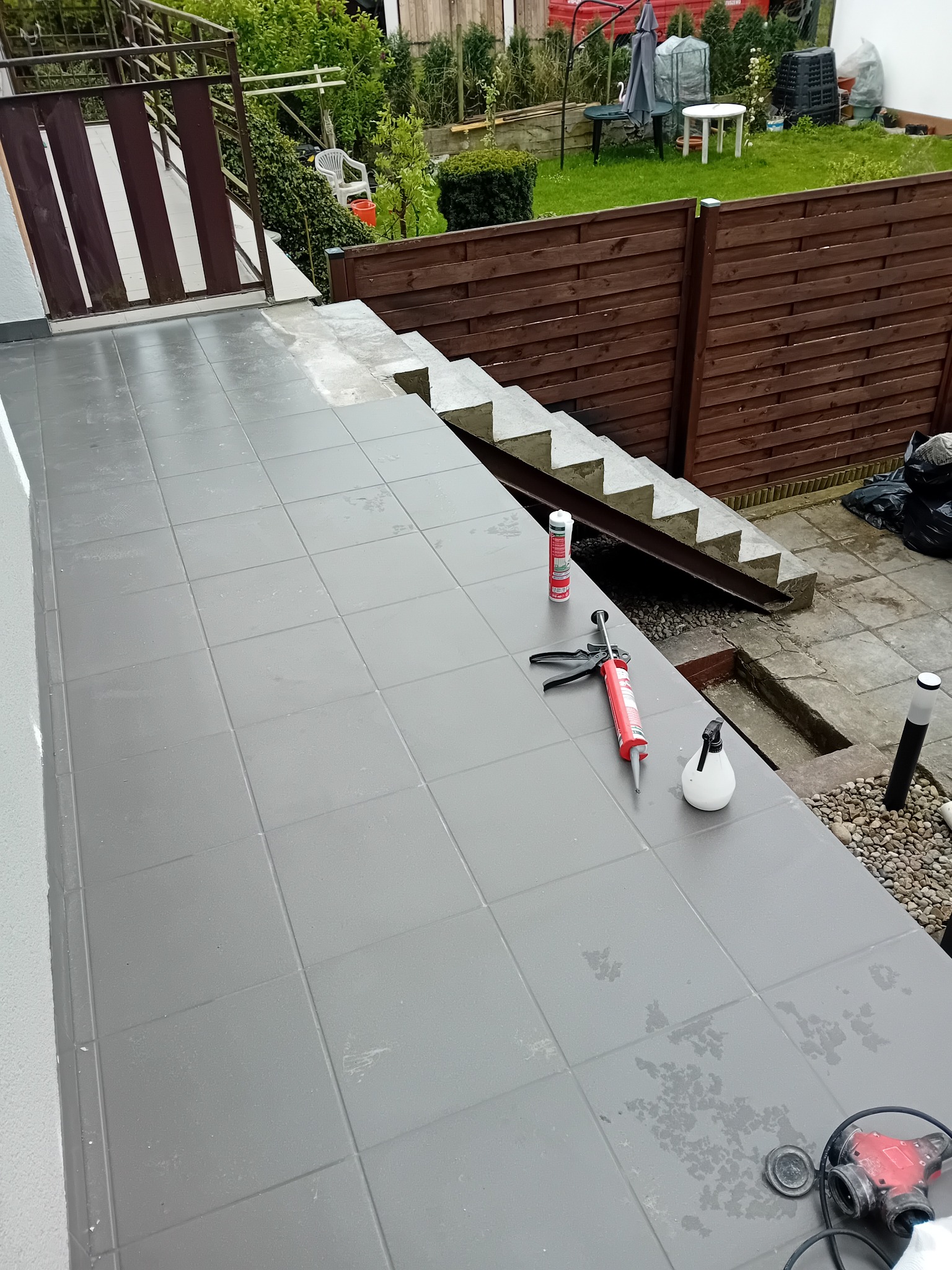 Szare płytki na tarasie w trakcie fugowania, widoczne narzędzia: pistolet do silikonu, pojemnik z płynem do czyszczenia, fuga w tubie, w tle schody i ogród.