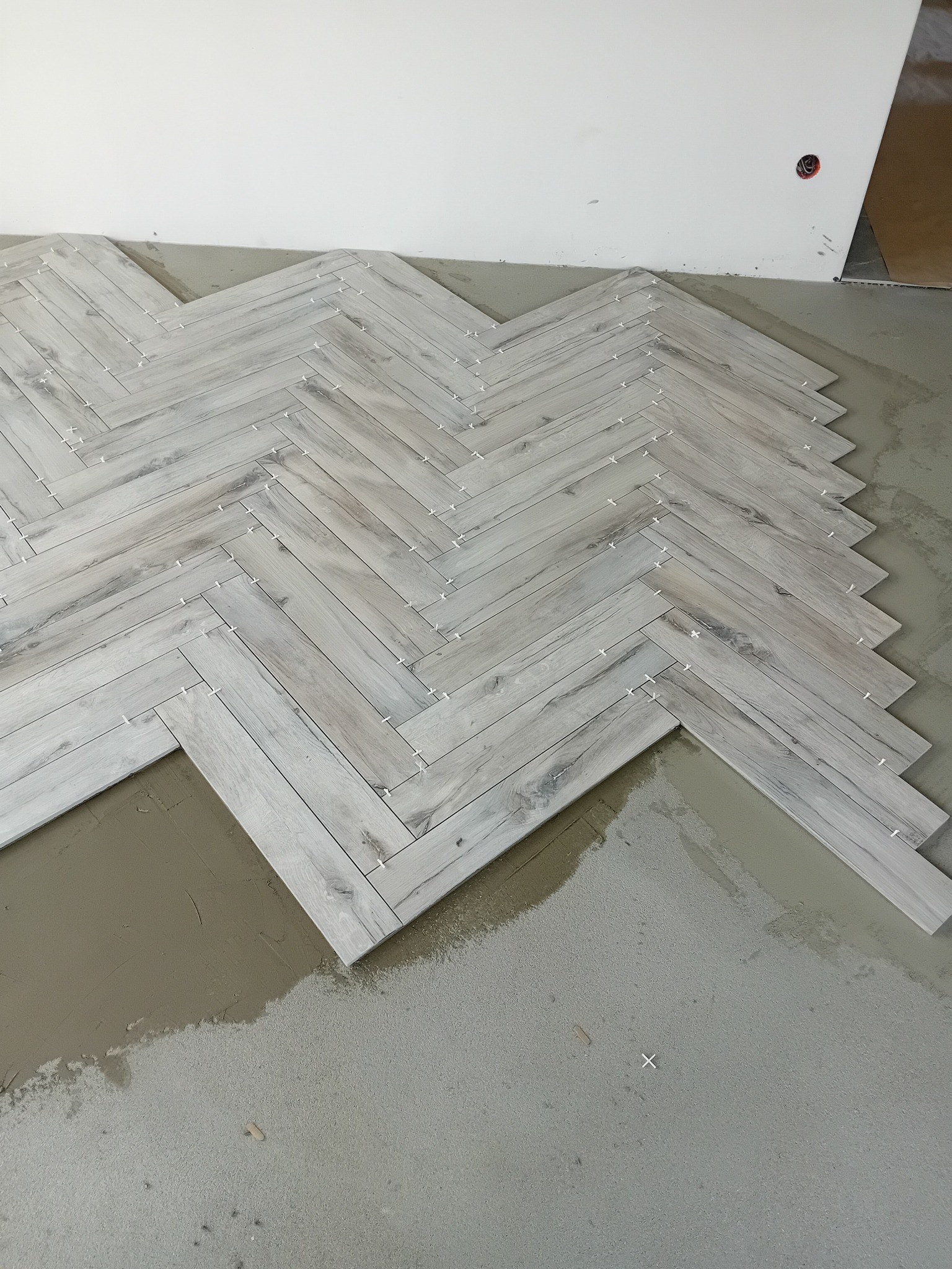 Układanie jasnoszarych płytek imitujących drewno w jodełkę, widoczne krzyżyki dystansowe i świeży klej na podłodze.