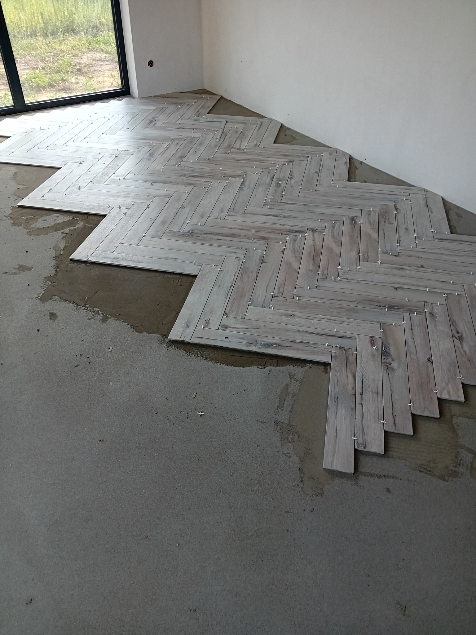 Układanie parkietu ceramicznego w jodełkę na podłodze w surowym stanie, widoczne krzyżyki dystansowe i świeży klej, duże okno w tle.