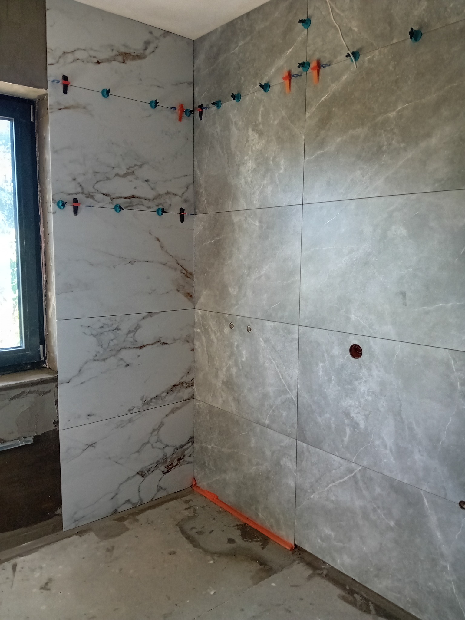 Ściana łazienki w trakcie układania płytek imitujących marmur, z użyciem systemu poziomowania płytek (klipsy i kliny), widoczne otwory pod instalacje.