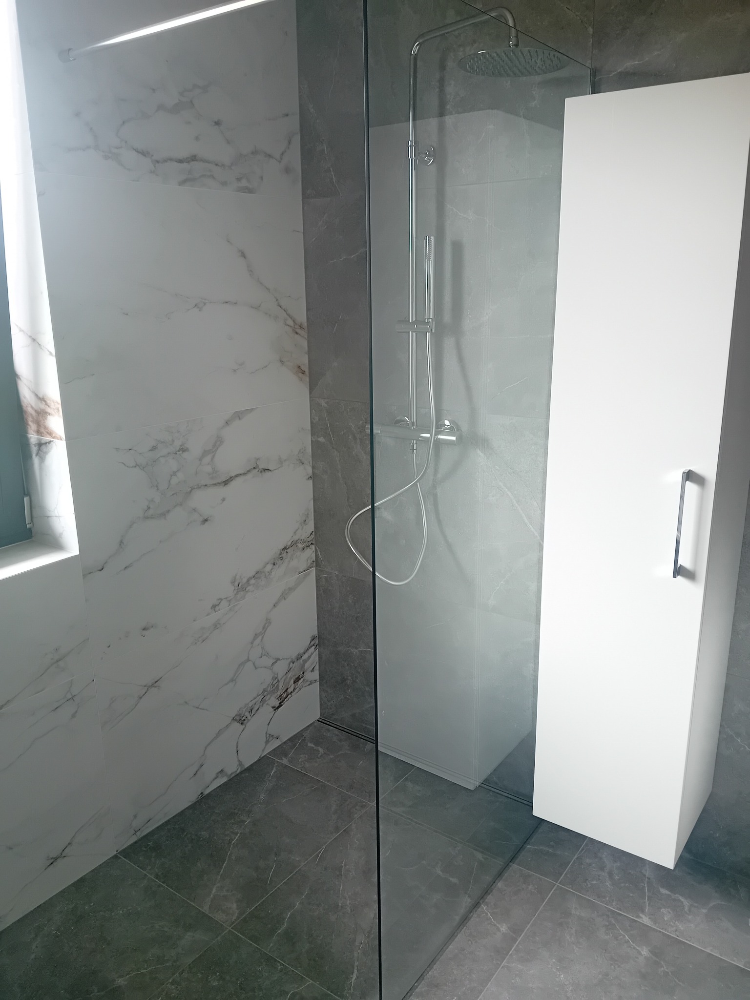 Nowoczesna łazienka z marmurowymi płytkami na ścianach, szarą podłogą, szklaną kabiną prysznicową z deszczownicą i wysoką białą szafką z chromowanym uchwytem.