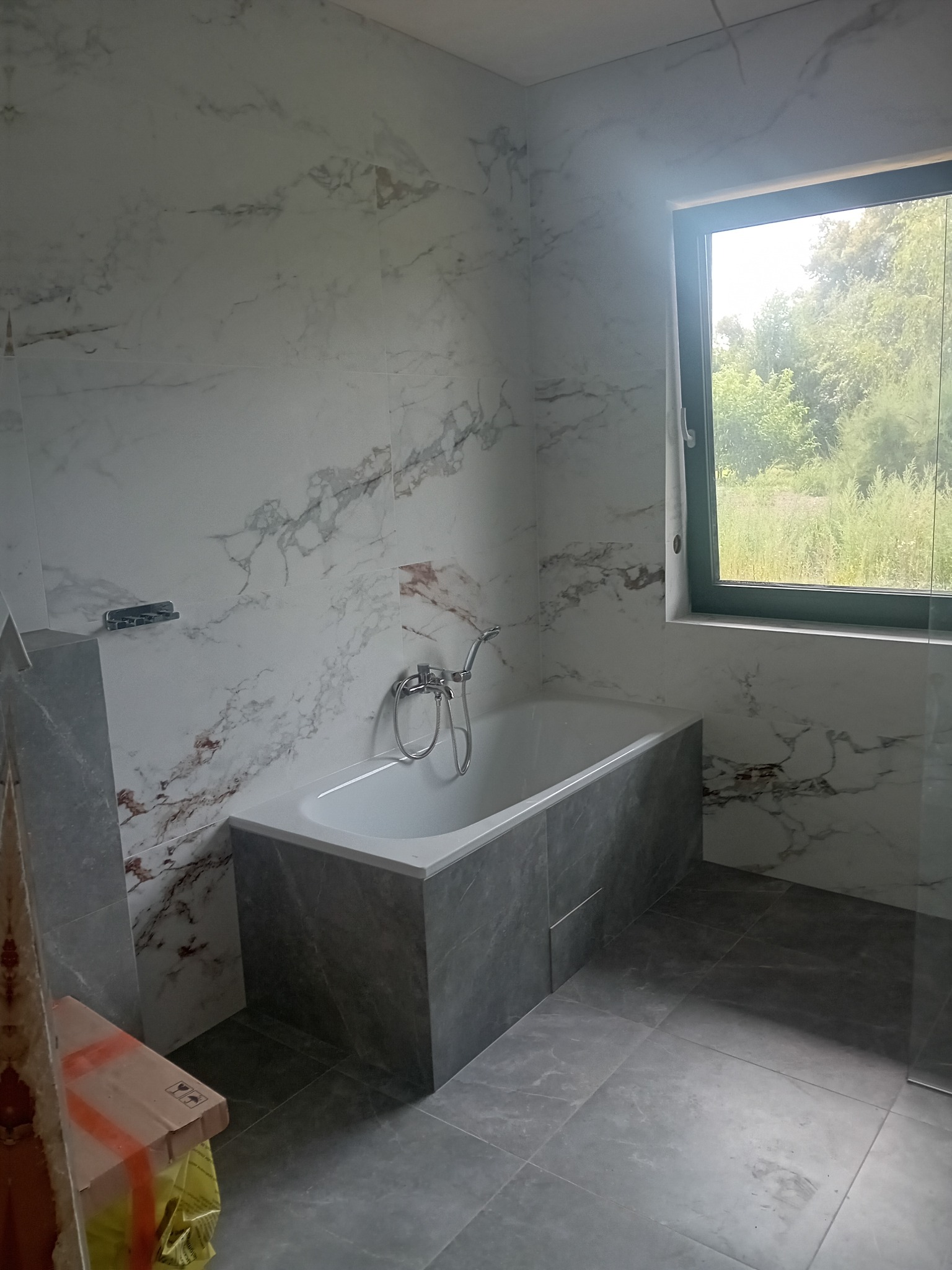 Wykończona łazienka z wanną w zabudowie z szarych płytek imitujących kamień, ściany wyłożone białymi płytkami z marmurkowym wzorem, widok na zieleń za oknem.