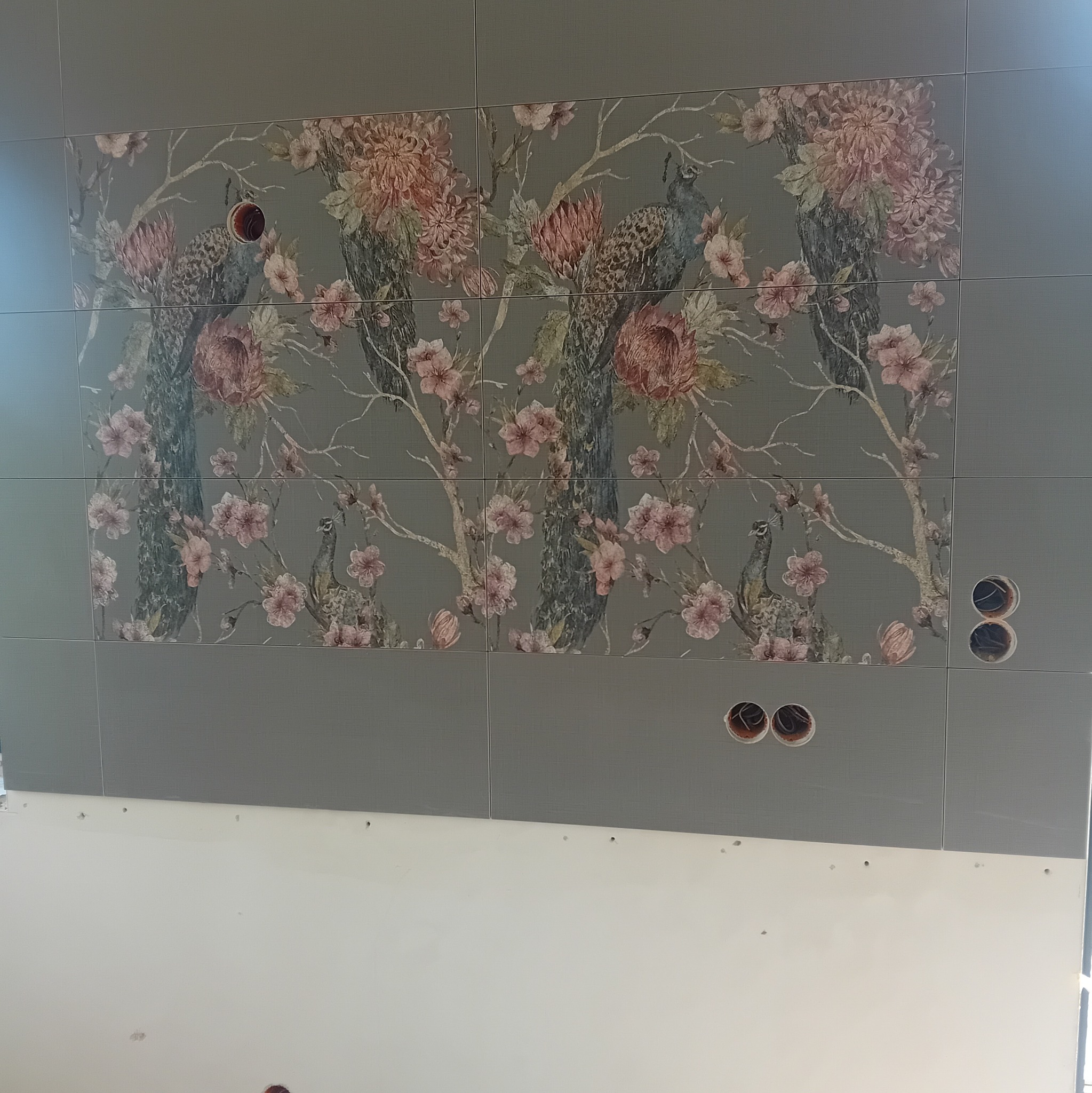 Ściana wyłożona szarymi płytkami z motywem kwiatowym i pawiami, widoczne otwory pod gniazdka elektryczne, dolna część ściany przygotowana do malowania.