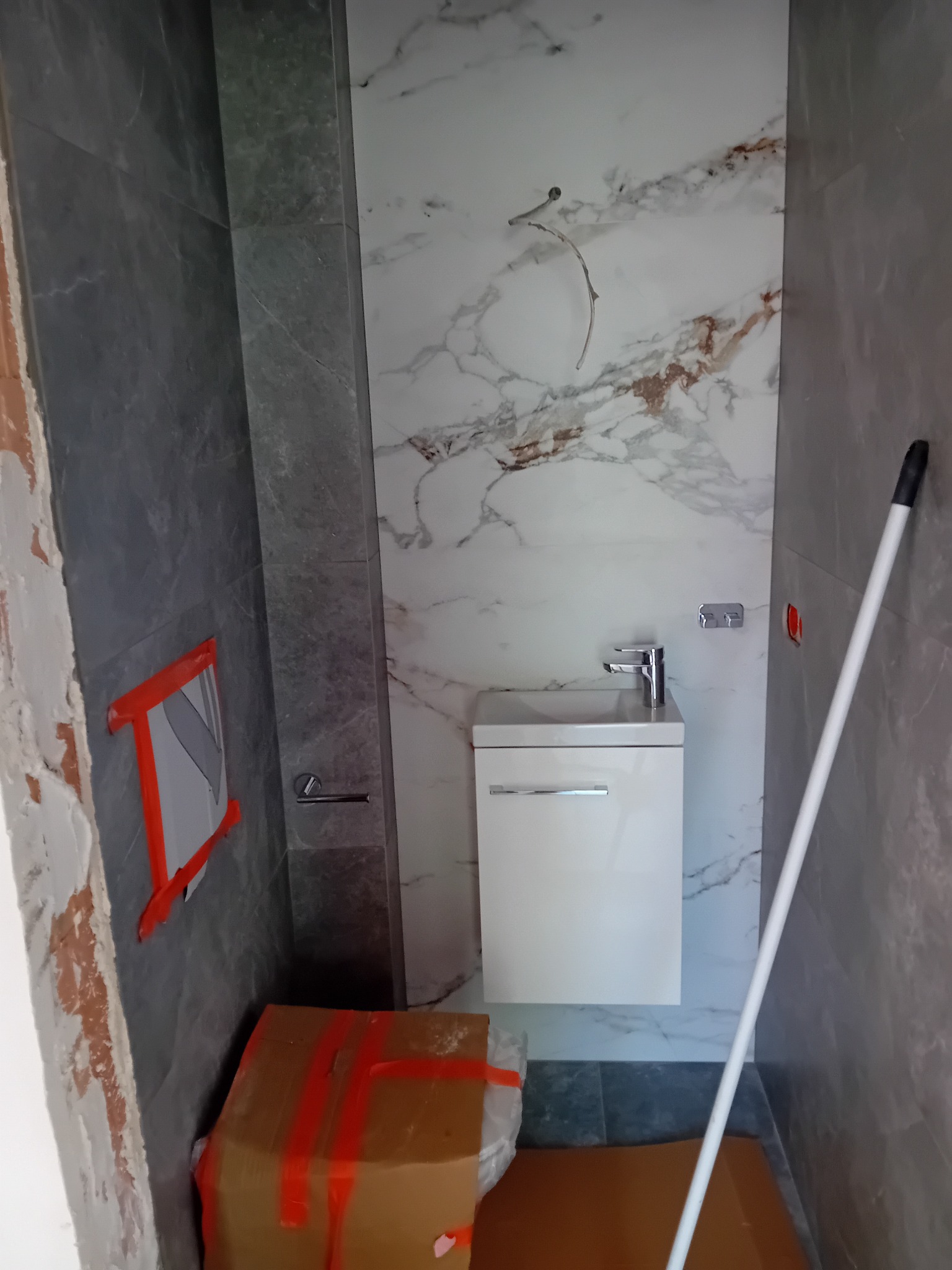 Wnętrze łazienki w trakcie remontu, widoczne płytki imitujące marmur na ścianie, zamontowana biała szafka z umywalką, częściowo wykończona ściana z szarych płytek, karton zabezpieczony taśmą...