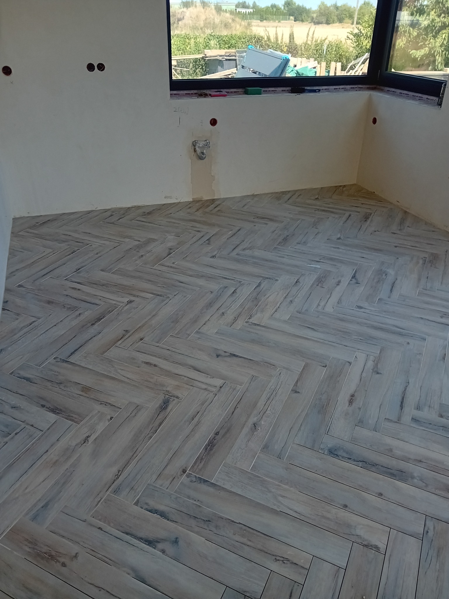 Ułożona podłoga z płytek imitujących drewno w jodełkę, widoczne niedokończone ściany z instalacjami elektrycznymi i okno z widokiem na zewnątrz.