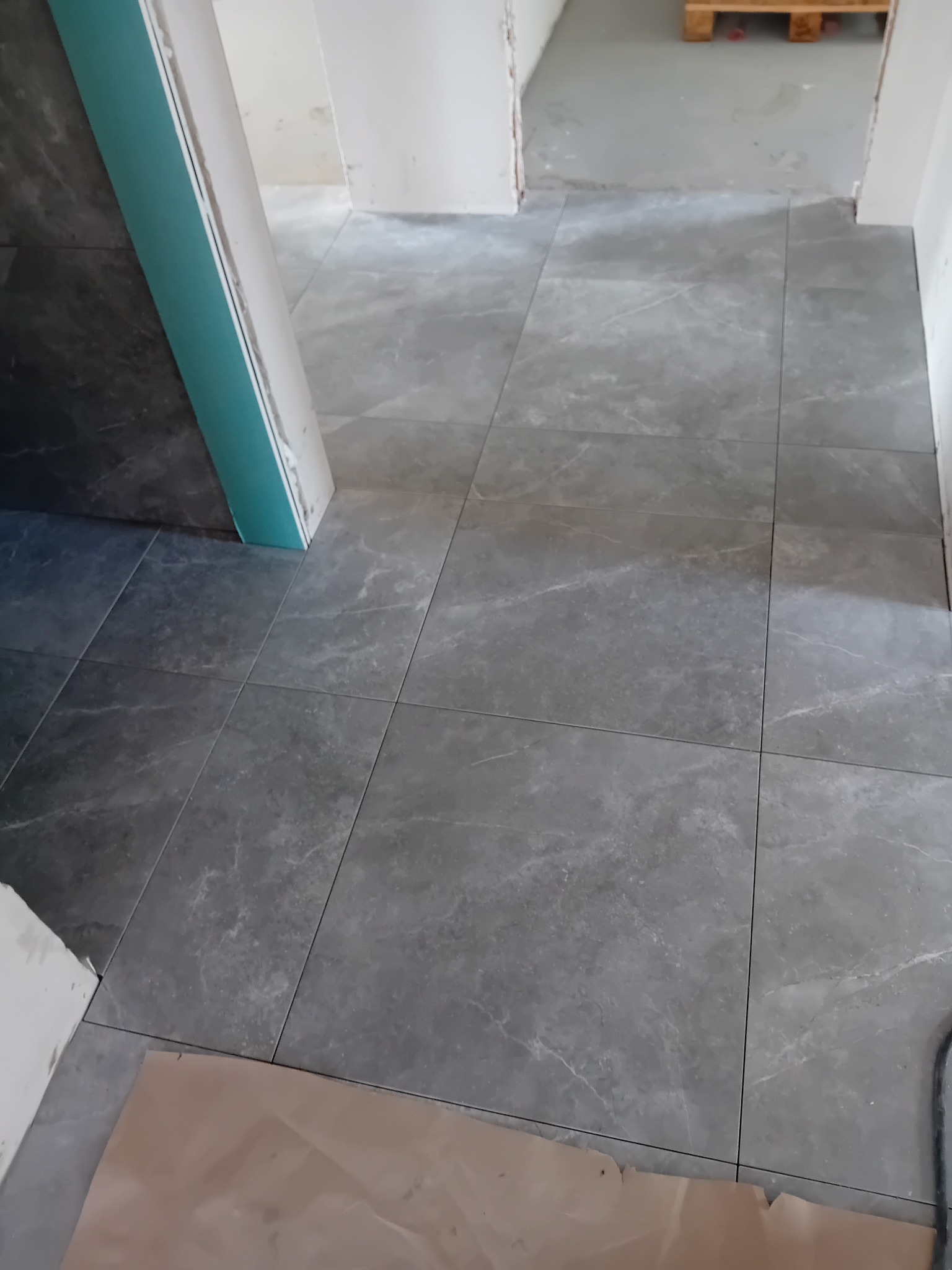 Ułożona szara, marmurkowana podłoga z płytek w trakcie remontu, widoczna paleta w tle i zabezpieczenie podłogi kartonem.