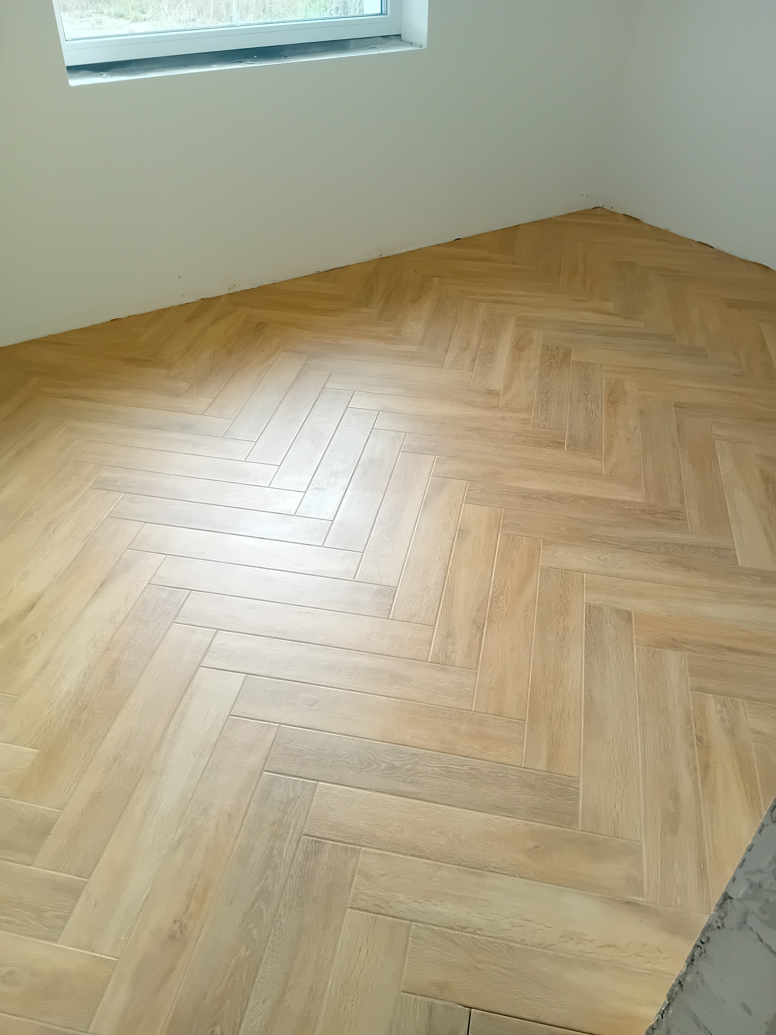 Ukończona podłoga z paneli ułożonych w jodełkę w jasnym odcieniu drewna, widoczne niedociągnięcia przy ścianie, narożnik pokoju, okno w tle.