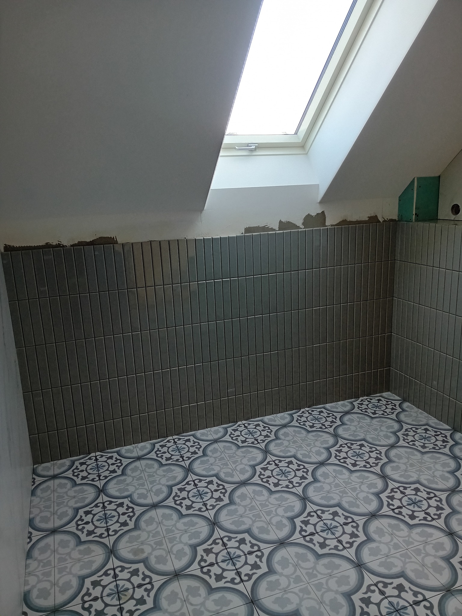Świeżo ułożone, pionowe, szare płytki na ścianie łazienki na poddaszu, z widocznym oknem dachowym i ozdobnymi płytkami podłogowymi w kwiatowe wzory. Częściowo widoczne ślady po kleju nad rzędem...