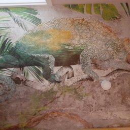 Rafał Jaroń Firma remontowo-wykończeniowa - Malowidło ścienne przedstawiające kameleona w tropikalnym otoczeniu, z widocznymi śladami pędzla i teksturą imitującą naturalny kamień.