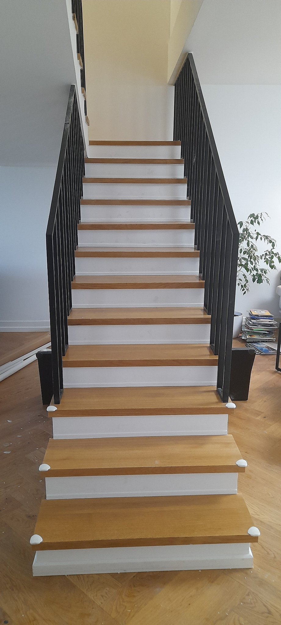 Schody drewniane z białymi podstopnicami i czarną metalową balustradą, zabezpieczone narożnikami ochronnymi, widok z dołu na wprost.