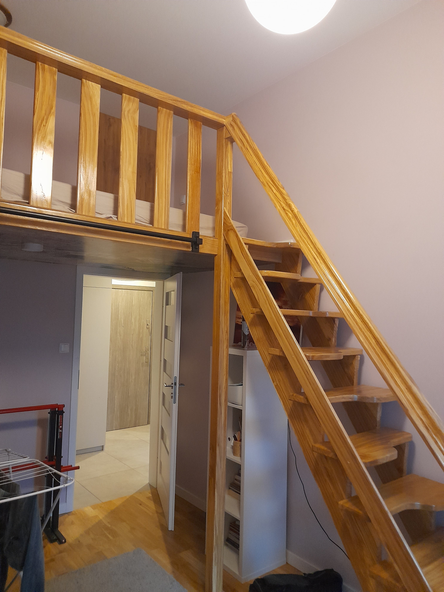 Drewniane schody prowadzące do antresoli z łóżkiem, widok z dołu, jasne drewno, białe ściany, minimalistyczny wystrój pokoju.