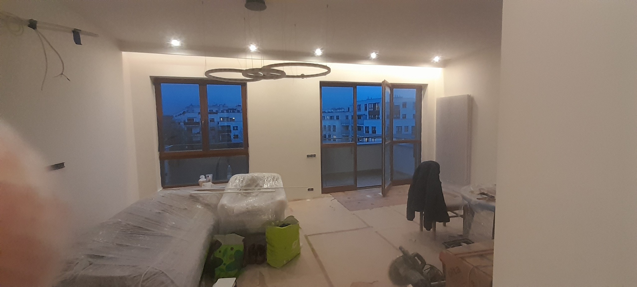 Wnętrze w trakcie remontu: okna z widokiem na miasto, zakryte meble, widoczne przewody elektryczne, podłoga zabezpieczona kartonem, nowoczesna lampa sufitowa.