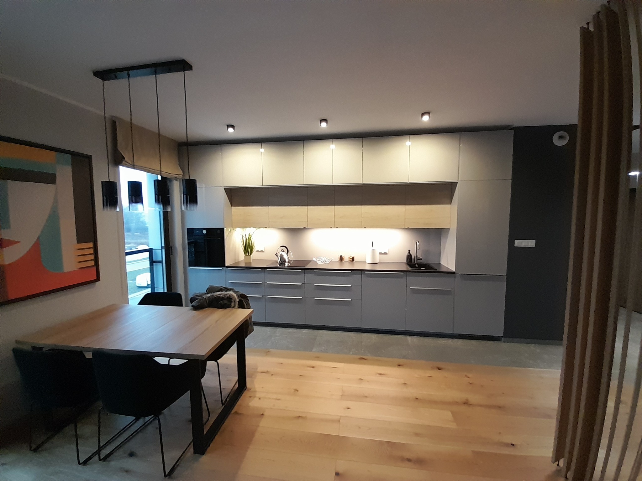 Nowoczesna kuchnia z szarymi szafkami, drewnianym blatem i podłogą. Nad stołem wiszą czarne lampy. Obraz na ścianie dodaje koloru. Minimalistyczny design.