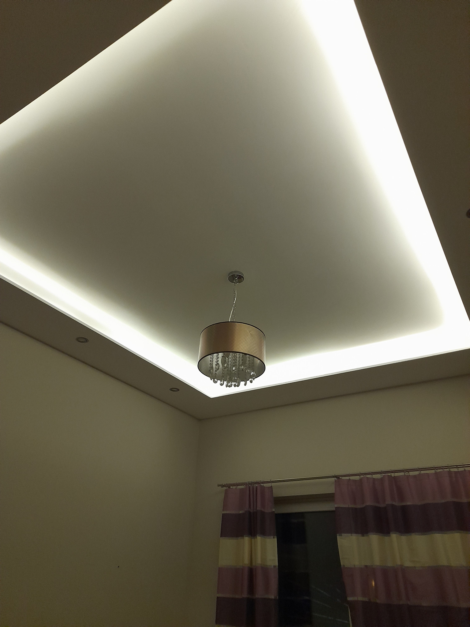 Sufit podwieszany z oświetleniem LED w kształcie prostokąta, centralnie umieszczony żyrandol z kryształkami, widoczne fragmenty ściany i zasłon w pokoju.