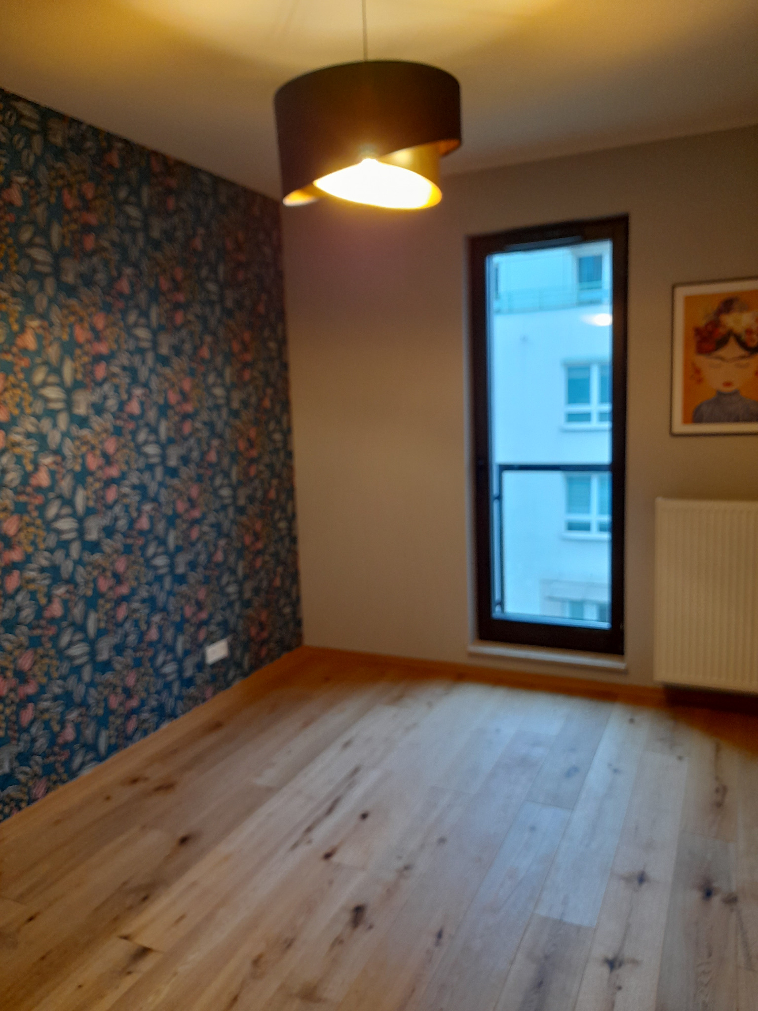 Pokój w Warszawie po renowacji, z drewnianą podłogą, ścianą pokrytą tapetą w kwiaty i nowoczesną lampą sufitową.