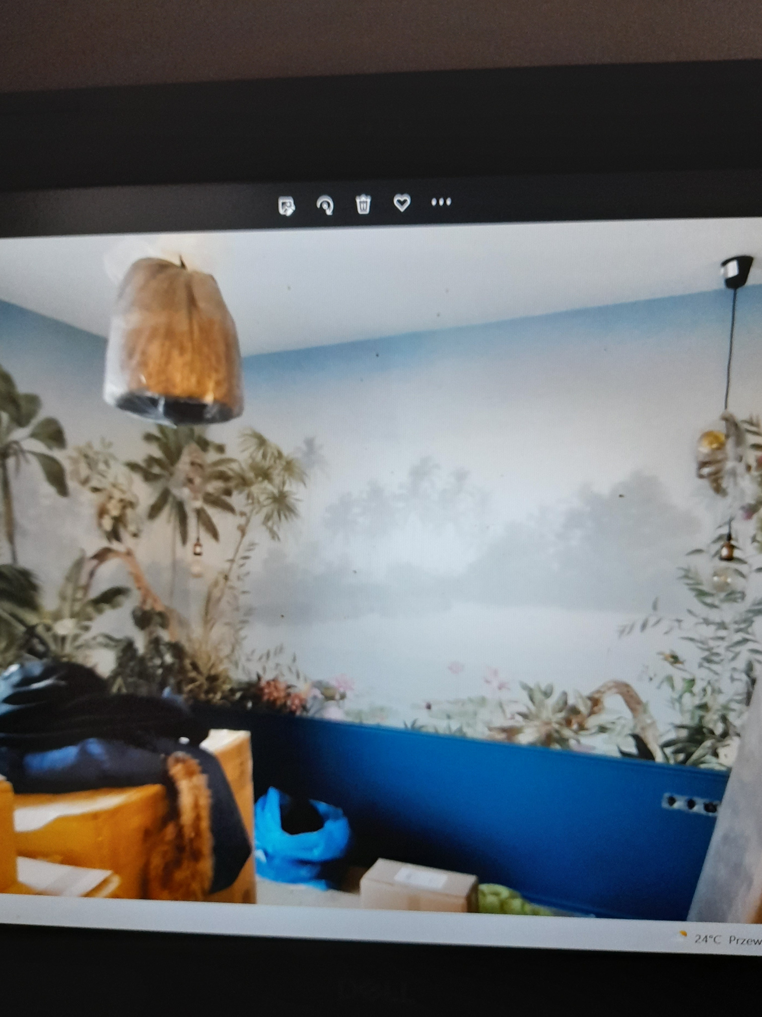 Pokój z fototapetą przedstawiającą tropikalny krajobraz, niebieski panel ścienny i wiszące lampy z abażurami, widok z góry na bałagan na pierwszym planie.