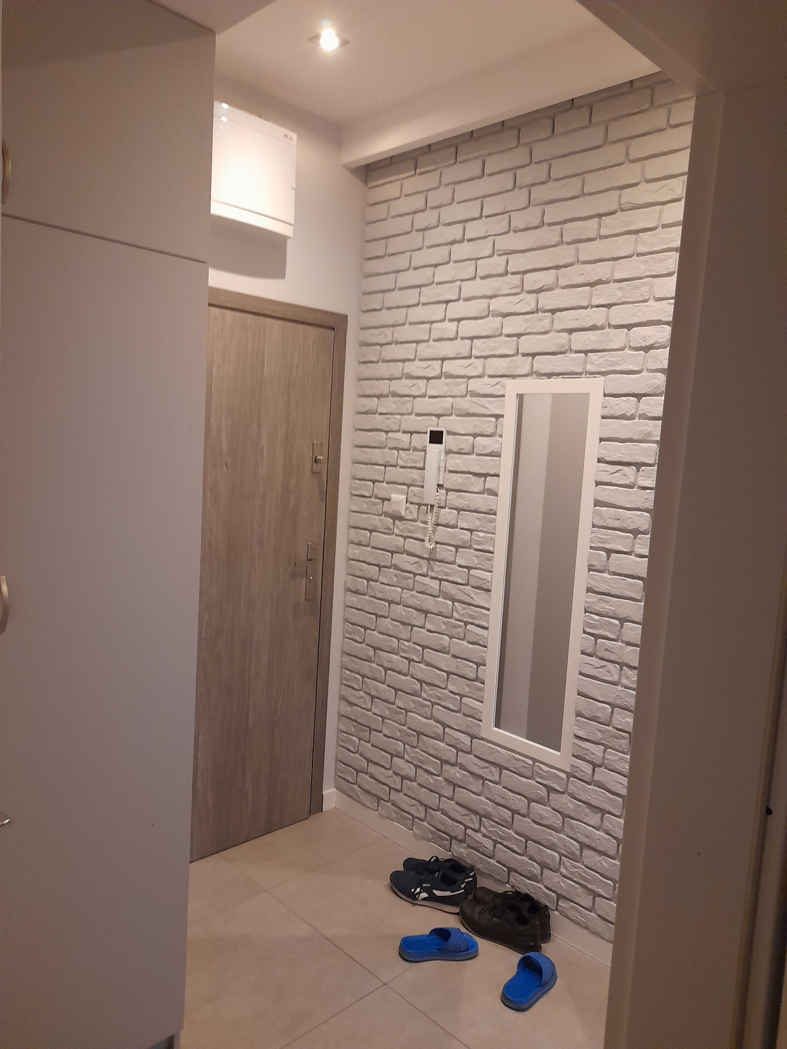 Przedpokój z białą cegłą dekoracyjną na ścianie, lustro w białej ramie, drzwi wejściowe w odcieniach szarości i beżu, oraz buty i klapki na podłodze.