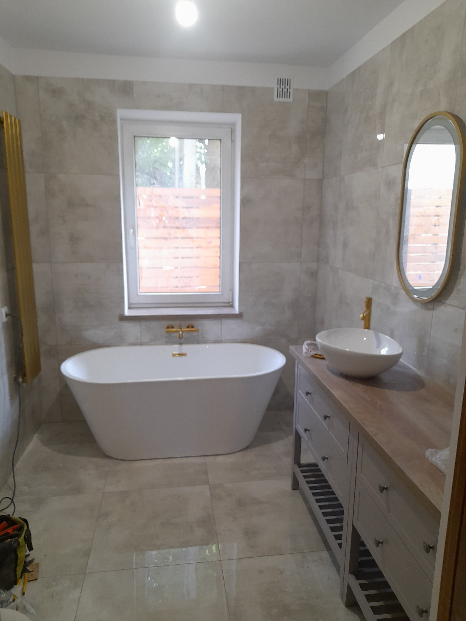 Nowoczesna łazienka z wanną wolnostojącą, umywalką nablatową na drewnianym blacie, szarą płytką imitującą kamień i złotymi akcentami w postaci baterii i grzejnika.