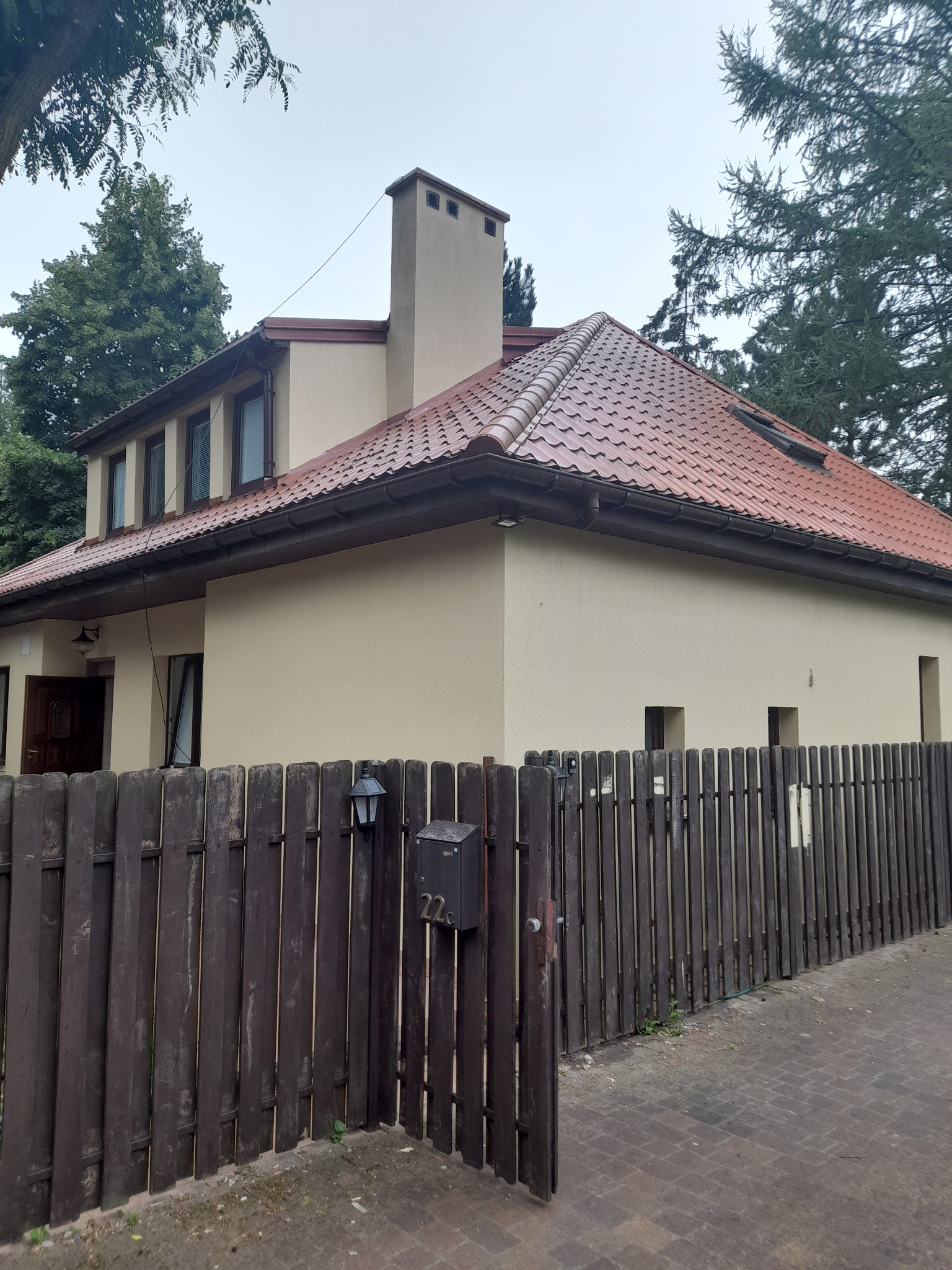 Dom jednorodzinny z beżową elewacją i czerwoną dachówką, widziany zza drewnianego płotu z numerem 22c na skrzynce pocztowej.