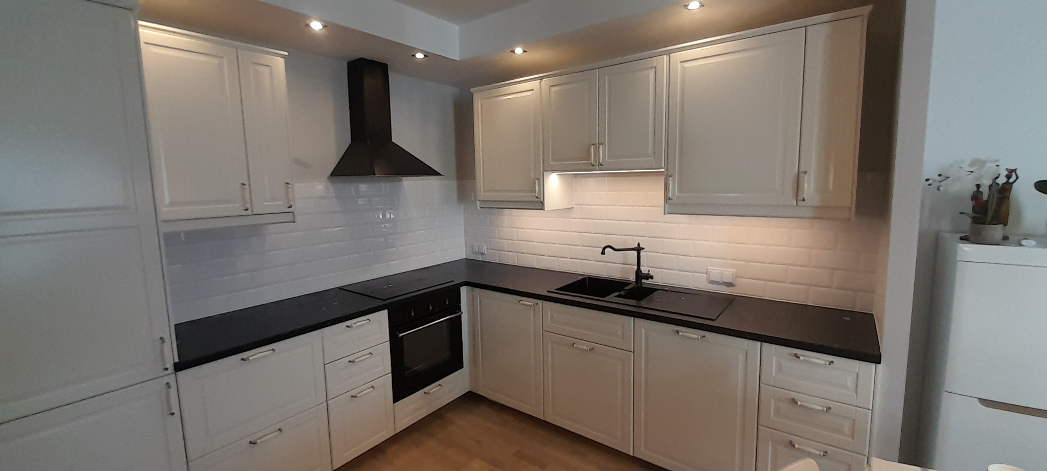 Nowoczesna kuchnia z białymi szafkami w stylu angielskim, czarnym blatem i czarną armaturą, wykończona białą cegiełką na ścianie.