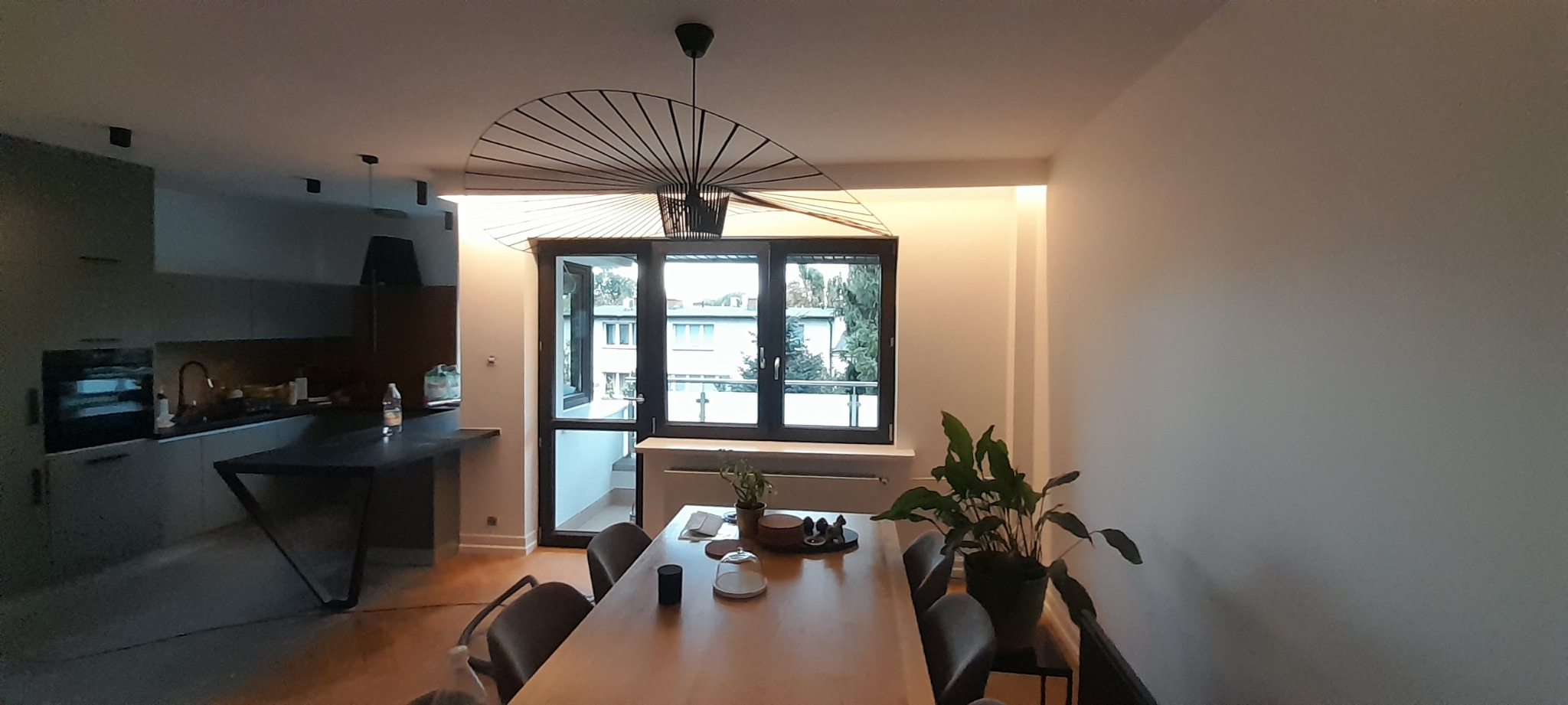 Nowoczesna jadalnia z drewnianym stołem, szarymi krzesłami, designerską lampą nad stołem i widokiem na zieleń za oknem balkonowym.