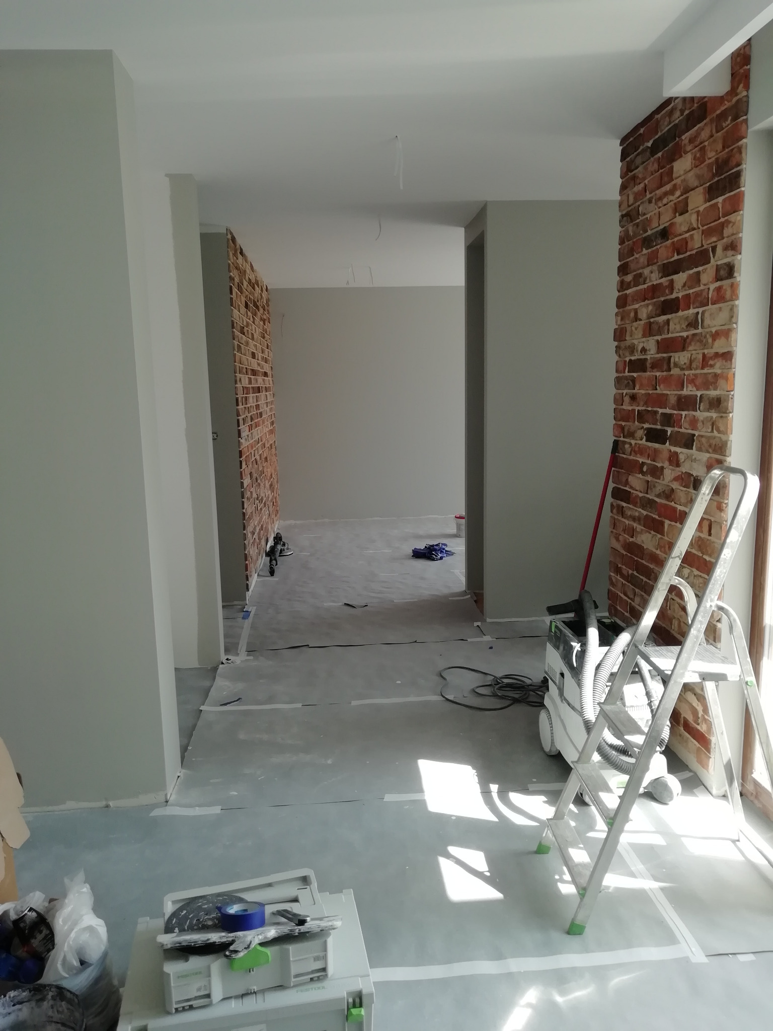 Wnętrze w trakcie prac wykończeniowych: szare ściany, fragment ściany z cegły dekoracyjnej, zabezpieczona podłoga, drabina i odkurzacz przemysłowy Festool.