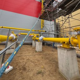 Yurii Ospishchev - Perfect Welding - Żółte rury gazowe z zaworami, wsparte na betonowych postumentach, na tle budynku z szaro-czerwonymi panelami i rusztowania. Prace instalacyjne w toku.