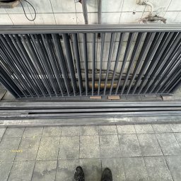 Yurii Ospishchev - Perfect Welding - Szara, metalowa konstrukcja kratowa z prętów, leżąca na podłodze z płytek, widziana z góry, z fragmentem stopy w kadrze. Elementy konstrukcyjne w tle.