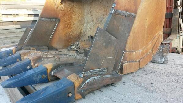 Naprawione zęby łyżki koparki z widocznymi spawami, ukazujące wzmocnienie konstrukcji. Detal z bliska, szaro-niebieskie zęby, brązowa łyżka, surowy beton w tle.