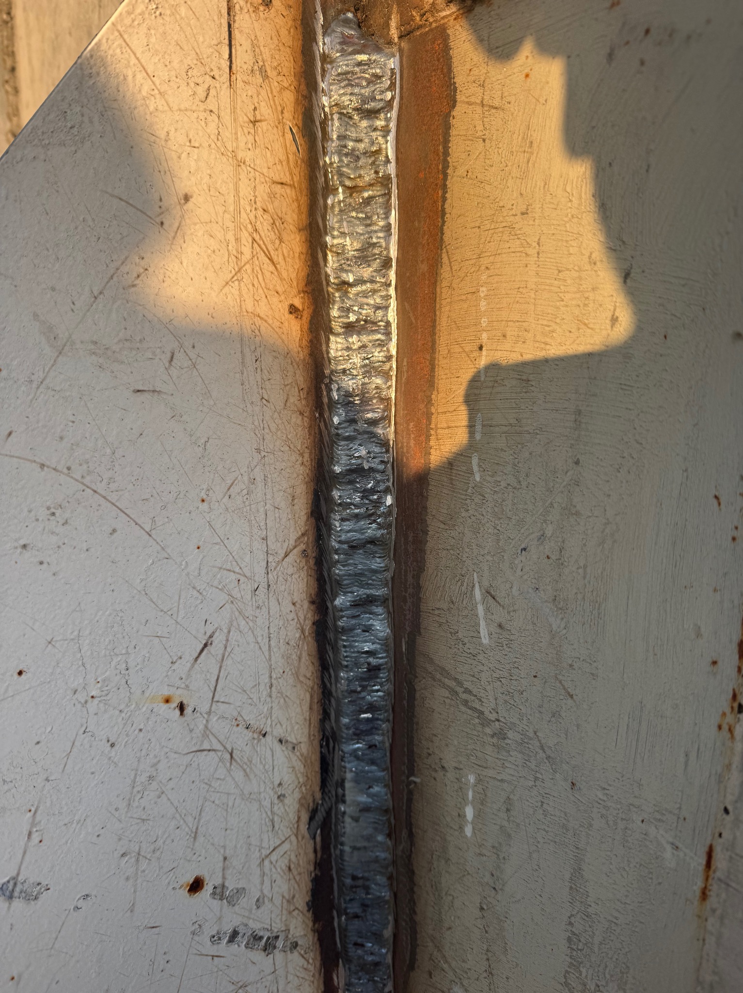 Spoina łącząca dwie metalowe płyty, widoczna z bliska, z delikatnym cieniem osoby w prawym górnym rogu. Szlifowana powierzchnia metalu.