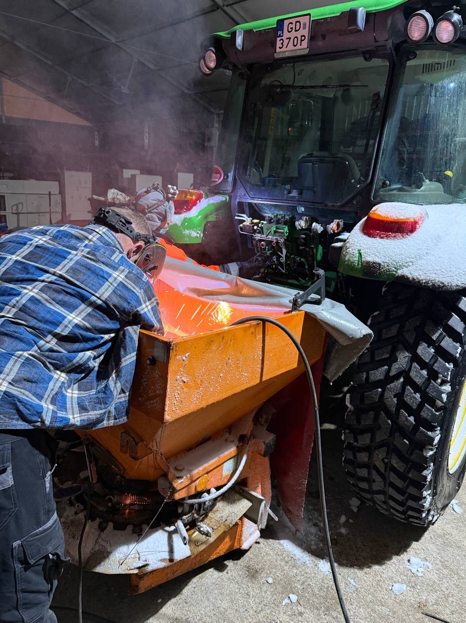 Prace przy pomarańczowym odśnieżaczu ciągnika: osoba w masce ochronnej spawa element, iskry, śnieg na oponach i urządzeniu. Warsztatowa sceneria.