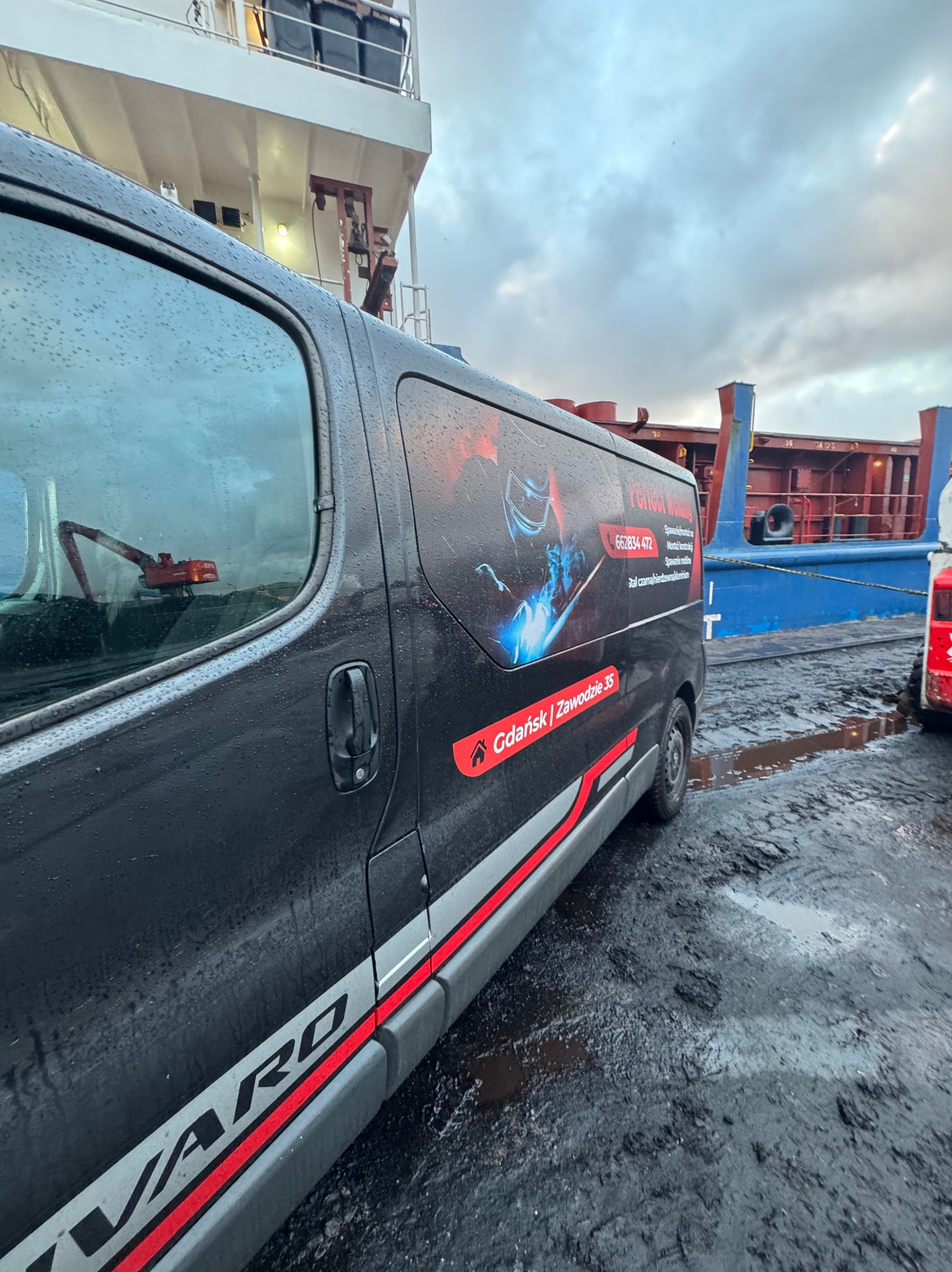 Czarny bus z grafiką spawacza i numerem telefonu na tle statku. Widoczne błoto na ziemi i krople deszczu na karoserii. Gdańsk, Zawodzie 35.