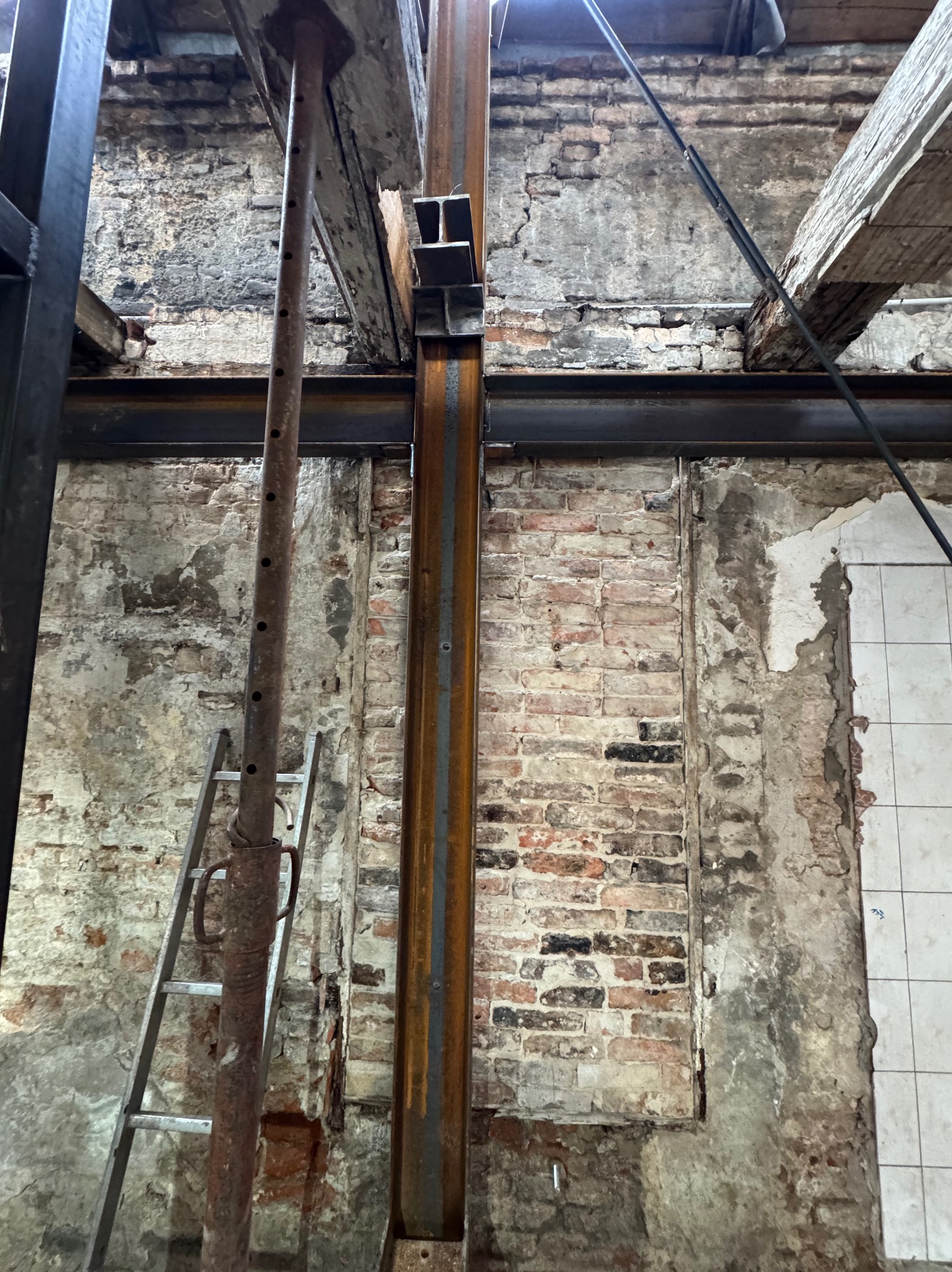Wzmocnienie konstrukcji budynku: stalowe belki i podpory na tle ceglanej ściany z fragmentami płytek. Widoczna drabina i elementy drewniane.