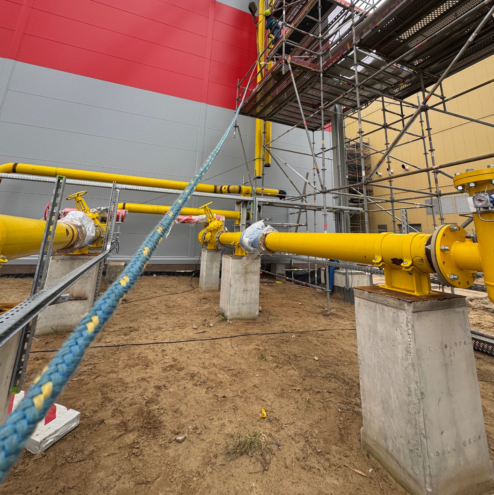 Żółte rury gazowe z zaworami, wsparte na betonowych postumentach, na tle budynku z szaro-czerwonymi panelami i rusztowania. Prace instalacyjne w toku.