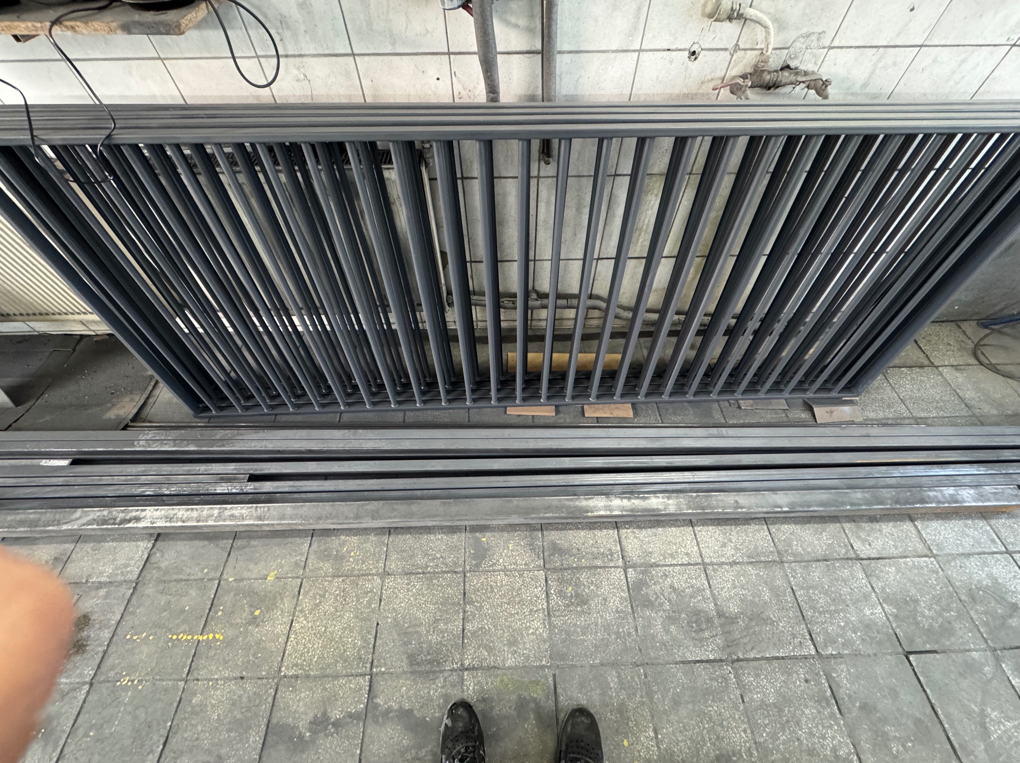 Szara, metalowa konstrukcja kratowa z prętów, leżąca na podłodze z płytek, widziana z góry, z fragmentem stopy w kadrze. Elementy konstrukcyjne w tle.